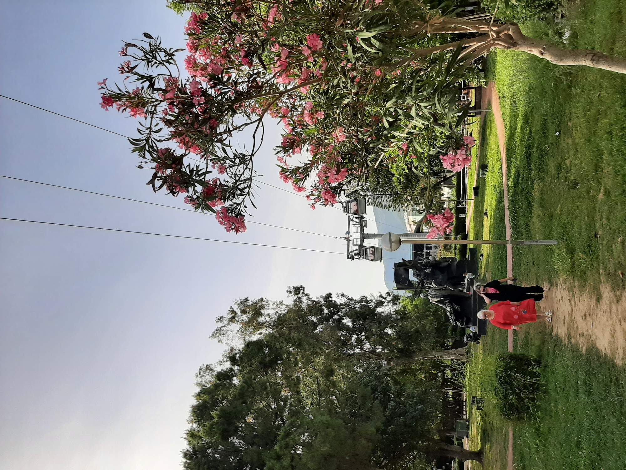 Two women walk along a dirt path in a park in Alanya, Turkey. One woman wears a red dress, and the other wears black clothing. They walk past a dark monument. A cable car travels overhead. Trees with pink flowers and leafy green trees flank the path. Grass and sky make up the rest of the scene.