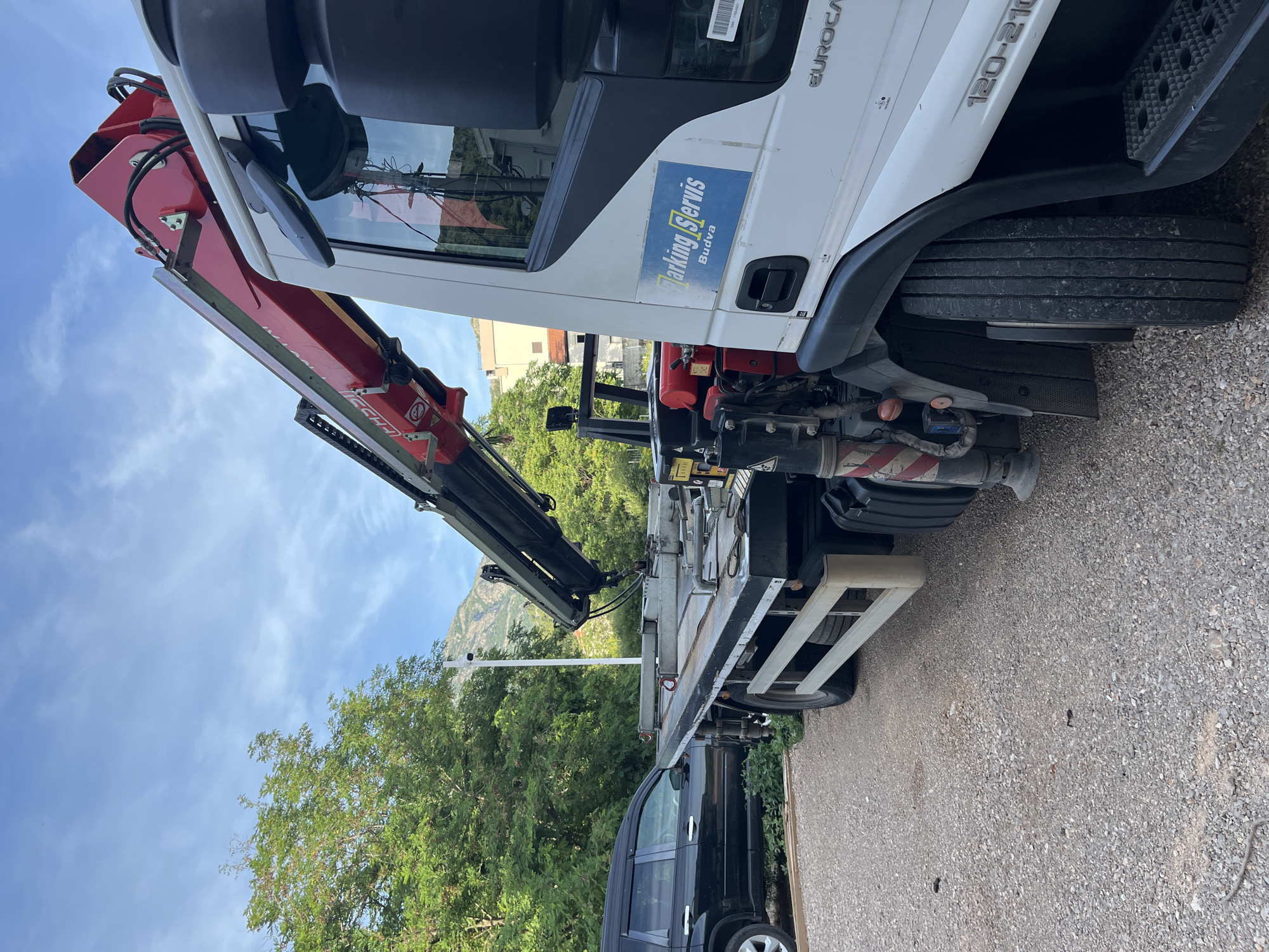 An exterior, eye-level, right-side shot captures a white flatbed truck equipped with a red crane. The truck is parked on a gravel surface. The crane's arm is extended upward and to the left. A black SUV is parked to the left of the truck. In the background, lush green trees and a light-colored building are visible. A mountainous landscape is also present behind the trees, under a blue sky with scattered white clouds. A sticker on the truck's door reads "Parking Servis Budva." The image appears to be taken in Budva, Montenegro.