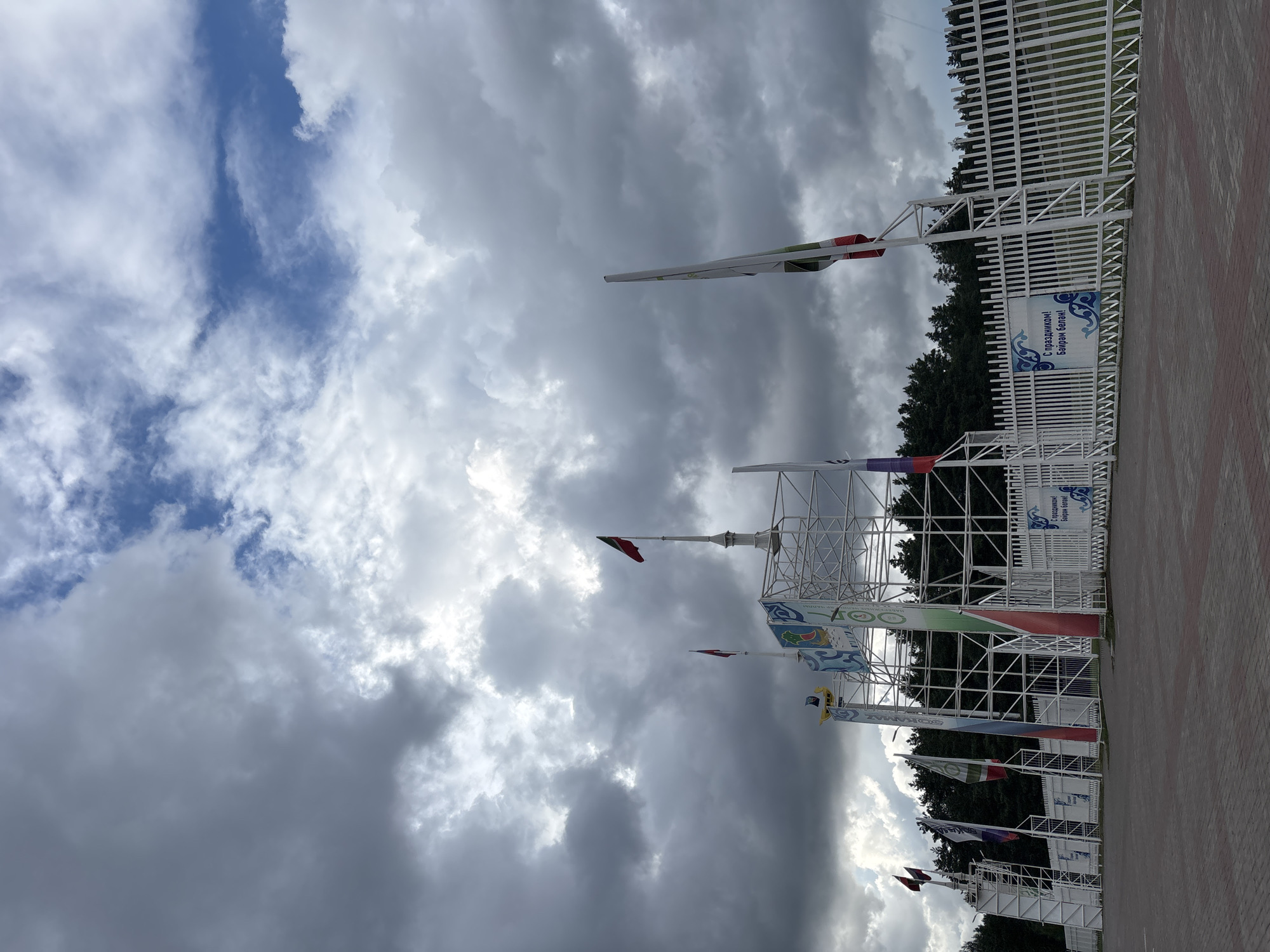 The image shows the entrance to the hippodrome in Naberezhnye Chelny, Russia. The entrance is a large white metal structure with flags flying from tall poles. The sky is cloudy with some patches of blue. In the foreground, there is a paved area and a white fence with banners. The banners have text in Cyrillic script. The hippodrome is surrounded by trees. There are no people visible in the image.