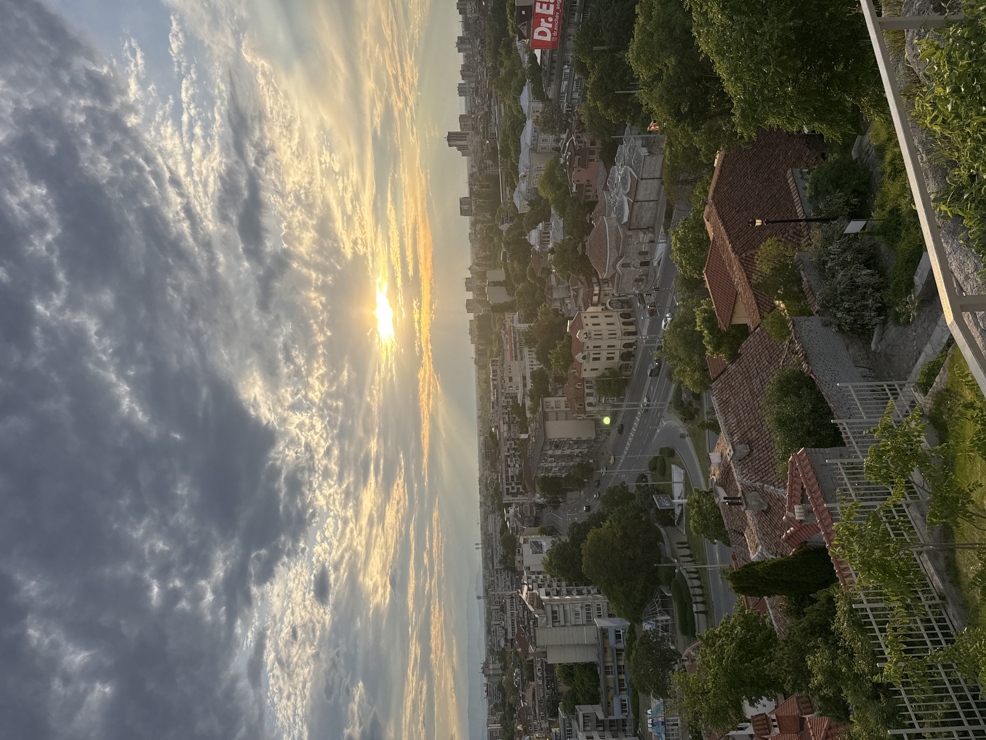 Captured from an elevated vantage point, the image showcases a city at sunset in Unknown, Unknown. The sky is predominantly cloudy, with the sun breaking through, casting rays across the horizon. Below, the cityscape is composed of buildings of various heights and architectural styles, interspersed with trees. A road runs through the city, showing vehicular movement. In the foreground, there's a glimpse of a white railing and greenery, suggesting the location is a balcony or terrace overlooking the city. A red sign with "Dr. El" is visible on a building further into the scene. The lighting is warm from the sunset, creating shadows and highlighting the textures of the buildings and trees.