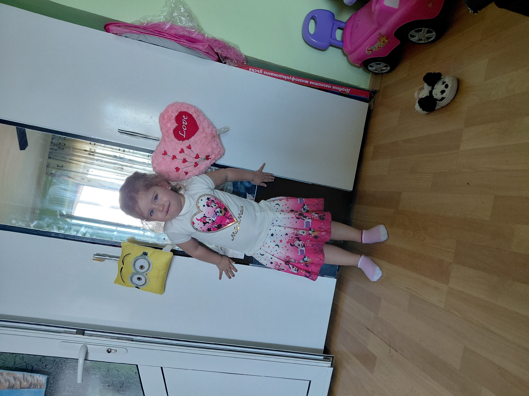 The image depicts a young girl standing in what appears to be a residential indoor setting, possibly a child's room or playroom. The girl is wearing a white t-shirt with a Minnie Mouse design and a pink skirt. She has blonde hair and is looking towards the camera with a neutral expression.
In the background, there is a large white wardrobe with mirrored doors. To the left of the girl, a yellow Minion plush toy is hanging on a hook. To her right, a pink heart-shaped pillow with the word "Love" written on it is also attached to the wardrobe.
The floor is made of light-colored wood laminate. On the floor, to the right of the girl, is a pink toy car with a purple handle and a white slipper with a black bow, resembling Mickey Mouse. The lighting suggests it is daytime, with light streaming in from a window, likely in the background, creating reflections on the wardrobe doors.
The image also shows a portion of a doorframe with a white door and a handle on the far left, and a pale green wall on the far right. There is a vertical piece of text in a Cyrillic alphabet visible on the right side of the wardrobe, possibly a product label or decoration.
The provided context mentions the city of Silistra, Bulgaria, suggesting this scene is located there. However, there are no specific visual cues within the image itself that confirm this location.