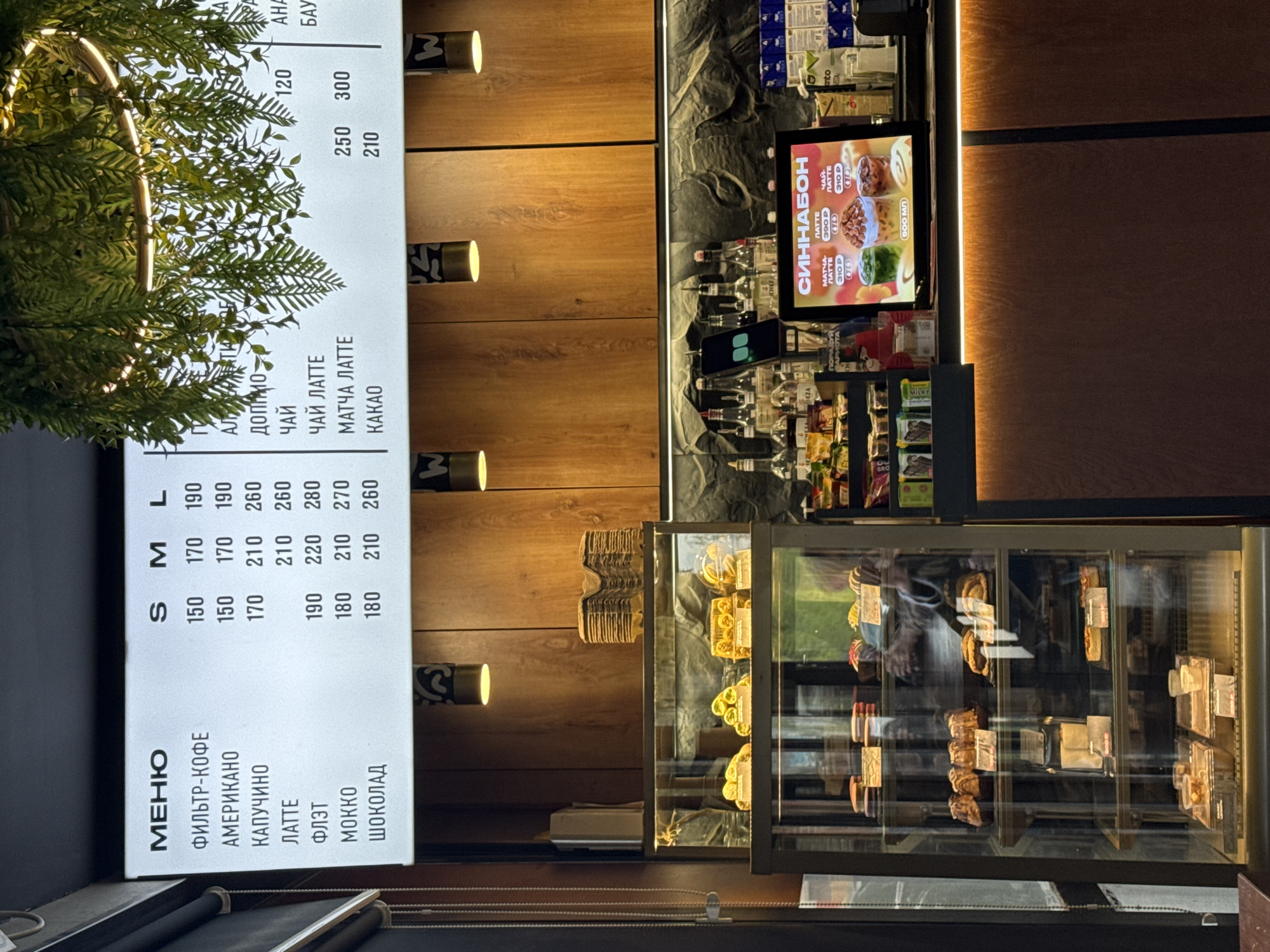 The image shows the interior of a coffee shop or café, located in Perm, Russia, based on the Cyrillic script in the menu. The photo captures several key elements:
1. **Menu:** A brightly lit board on the left displays the menu, offering various coffee options like "Filter Coffee," "Americano," "Cappuccino," "Latte," "Flat White," "Mocha," and "Chocolate," as well as tea, matcha latte, and cacao. Prices are listed in Russian rubles (presumably) in small, medium, and large sizes.
2. **Refrigerated Display Case:** Adjacent to the menu is a glass-fronted display case filled with pastries and desserts. The items are arranged on shelves, showcasing a variety of baked goods.
3. **Counter Area:** Above the refrigerated case, a counter area is partially visible, stocked with various beverages and snacks. A digital display shows a "Синабон" (Cinnabon) advertisement.
4. **Interior Design:** The interior features warm wood paneling and decorative lighting. Foliage has been incorporated in the display.
The scene presents a typical cafe ambiance, focusing on the point of sale and product display.