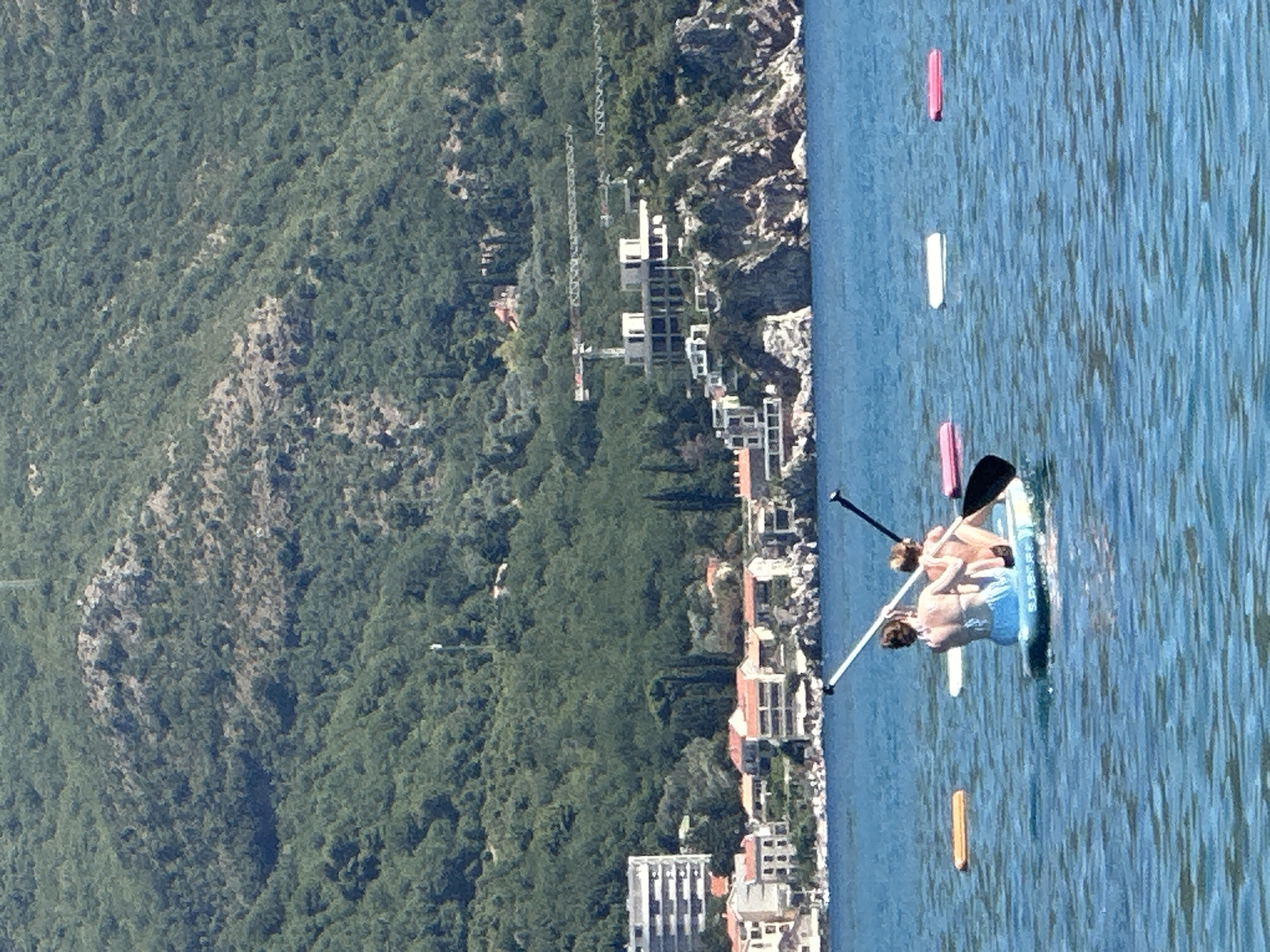 In Boreti, Montenegro, two individuals are paddleboarding on a bright, sunny day. The sea is calm and a vibrant blue. The paddleboarders, viewed from behind, are positioned in the foreground of the image. One individual is sitting on the paddleboard, wearing a blue bikini bottom, while the other is behind them, also seated. Both are holding paddles. In the background, a shoreline is visible with buildings ranging from multi-story structures to smaller houses with red roofs. Behind the buildings, a lush green, densely forested mountain slopes upwards. Several construction cranes are also visible on the mountainside, indicating ongoing development. Additionally, there are several colorful inflatable mats floating on the water in the mid-ground.
