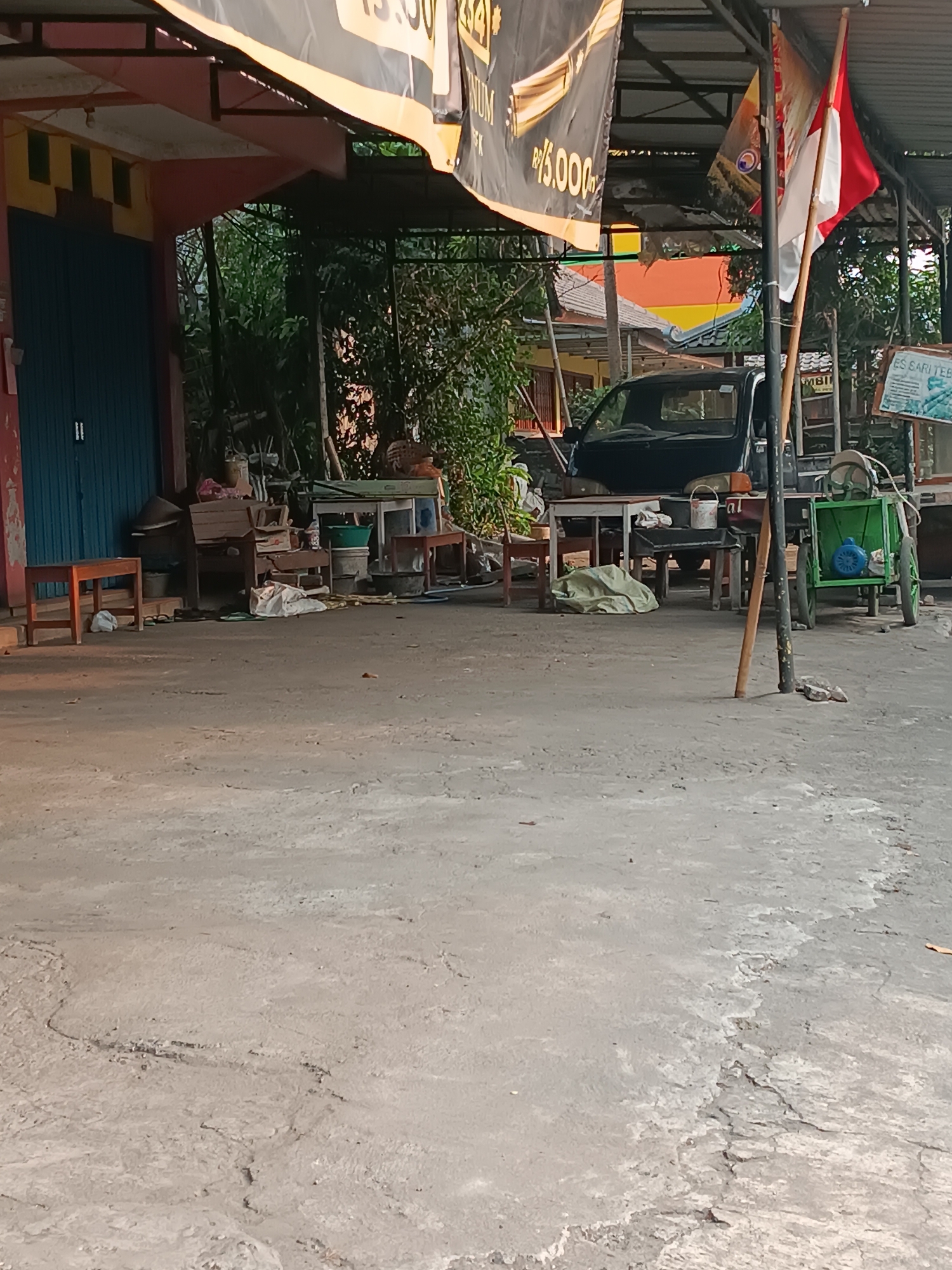 The image depicts an outdoor scene in Pleret, Indonesia, likely a roadside stall or small business. In the foreground, a cracked concrete ground is visible, with some leaves scattered on it. Further back, under a corrugated metal roof supported by metal poles, a blue roller-door shop front is to the left, with a small wooden stool in front of it. Adjacent to the shop, there are various items including wooden furniture, plastic buckets, a green cart with wheels, and a black pickup truck. A Indonesian flag, red and white, flies from a wooden pole in the center of the scene. Behind the structures, lush green trees and vegetation are present. A sign with the text "ES SARI TEH" and an image of tea is visible on the right. The overall impression is one of a functional, perhaps slightly cluttered, commercial area in a tropical environment.