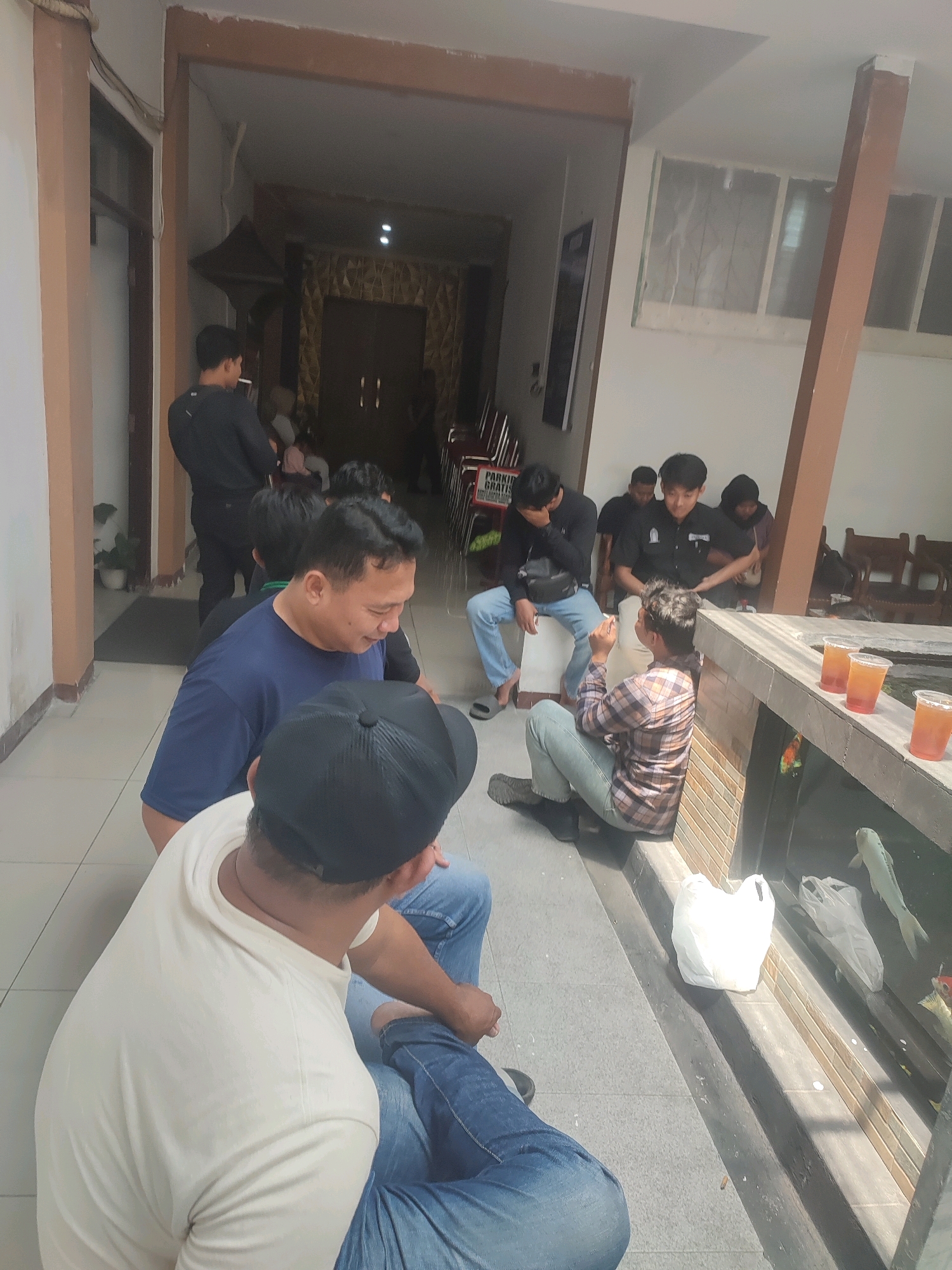 A group of approximately ten young men are gathered in an outdoor, covered walkway, possibly a waiting area or a communal space. The walkway has tiled flooring and is illuminated by overhead lights and natural light filtering in from an unseen area. To the right, there's a water feature or pond with at least two koi fish visible and several plastic cups containing amber-colored liquid. Two white plastic bags are also placed near the pond. Several of the men are seated on a low ledge or steps. One man in the foreground, facing away from the camera, wears a cream-colored t-shirt and blue jeans, with a black baseball cap. Another man, slightly to his left, wears a blue t-shirt and blue jeans. In the background, other men are seen standing and sitting, some are looking at their phones. The architecture suggests an Indonesian setting, and given the context of Nganjuk, it could be a public space, a residential complex, or a commercial establishment. The overall atmosphere appears casual and relaxed.