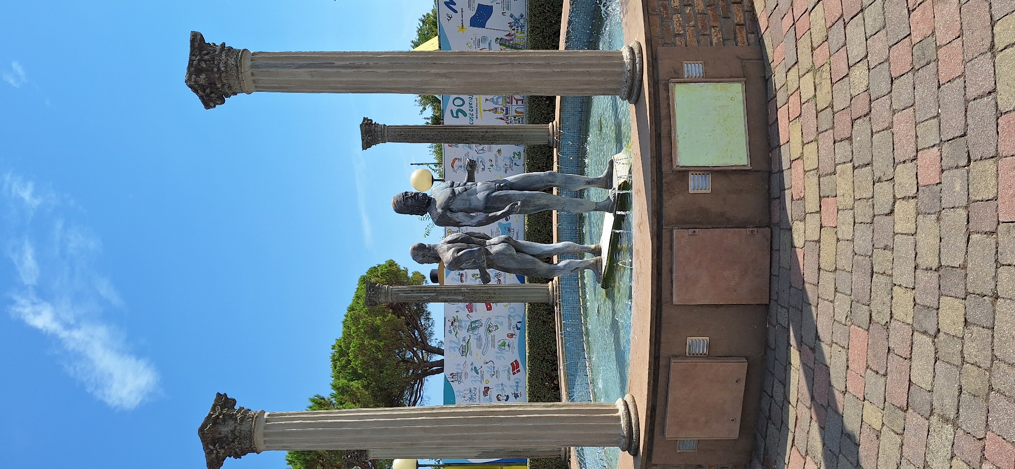 This is a full-frame, eye-level shot of a fountain with two male statues in Rimini, Italy. The fountain is located in front of a colorful mural depicting various scenes and numbers, likely educational or promotional in nature. The fountain itself is circular, with a brick-lined base. The two statues, rendered in a bronze or weathered metal material, appear to be historical or mythological figures. One statue stands with his back to the viewer, while the other faces partially towards the left. They are positioned on a white platform in the center of the fountain, surrounded by water with visible ripples and spray.
The fountain is framed by four large Corinthian columns, two on the left and two on the right, adding a classical architectural element to the scene. The columns are made of a light-colored stone and show some signs of age and wear. Behind the mural and the fountain, a lush green tree is visible, suggesting an outdoor setting. The sky above is a clear, vibrant blue with a few wispy white clouds.
The ground in front of the fountain is paved with a textured pattern of interlocking bricks in varying shades of red, yellow, and grey. The lighting suggests a bright, sunny day, casting distinct shadows on the brickwork. The overall scene evokes a sense of public space, possibly a park, plaza, or attraction in Rimini.