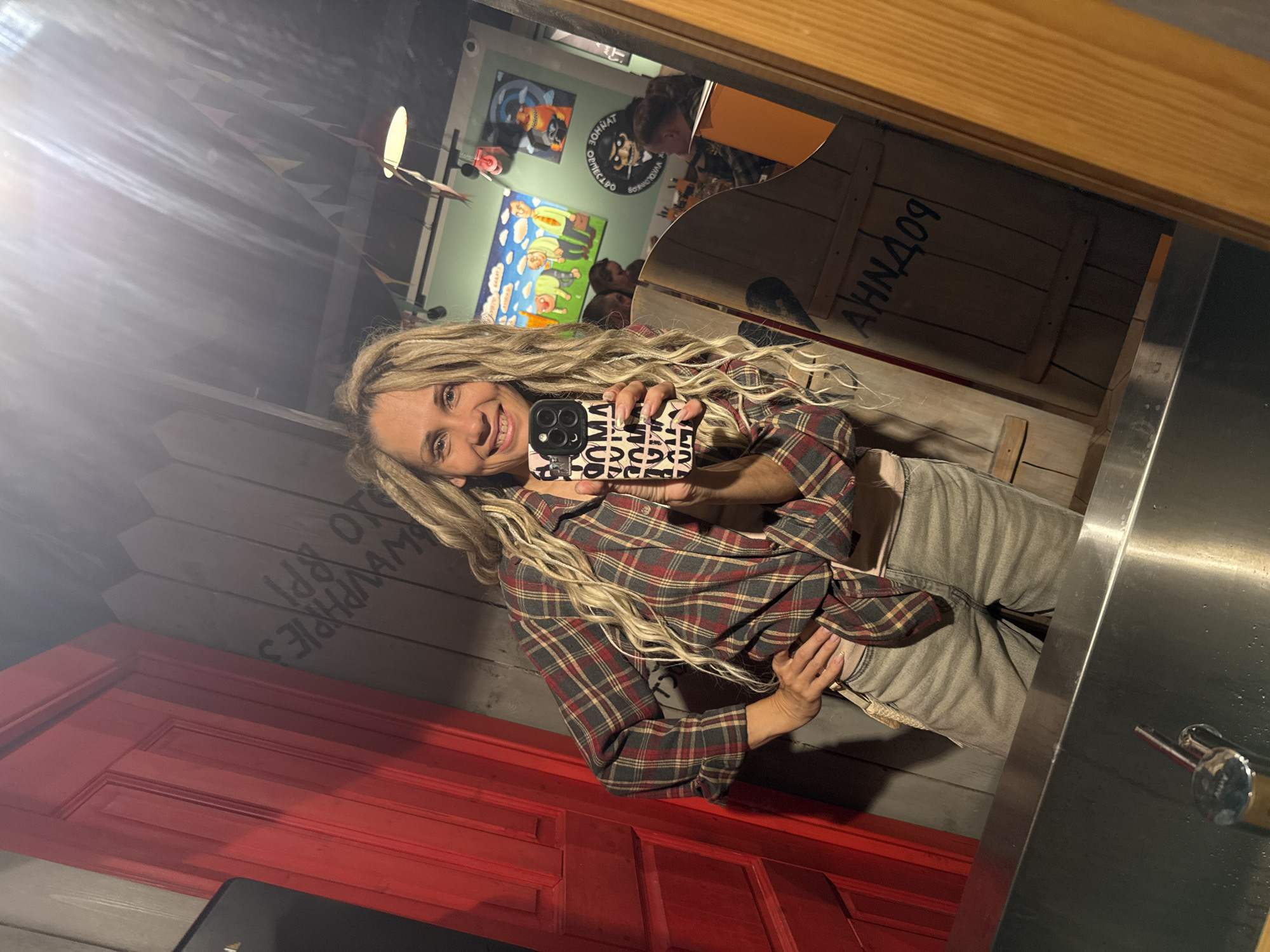 A woman with long, light-colored dreadlocks or braids is captured in a mirror selfie indoors. She is smiling broadly, holding a smartphone with a distinct black and white patterned case featuring repeating letters, possibly "LOVE." She is dressed in a plaid shirt with red, grey, and beige checks, and light grey pants.
The reflection reveals an eclectic interior, likely a casual restaurant or bar, in Заволжье, Russia. To the left of the reflected image, a bright red panel resembling a door is visible. To the right, a light green wall is adorned with several colorful, cartoonish paintings, including one depicting figures in a landscape and another showcasing a character with a beard and hat. Above these artworks, the Cyrillic text "ОБТОВНАЖ ЗОМНАТ" is visible.
Further right, a section of dark wooden paneling displays the reflected Cyrillic text "АНЧАОЯ." Below the red door, another wooden panel features partially visible, reflected text, appearing as "Sppl ЭТО & SYAHWVA AM," which combines Latin and Cyrillic scripts.
The setting appears to be lit by artificial light, and a bright light source in the upper left of the image creates a lens flare. A stainless steel counter or sink area is visible in the right foreground of the reflection. Blurry reflections of other people can be vaguely discerned in the background. The overall impression is a vibrant, informal atmosphere.