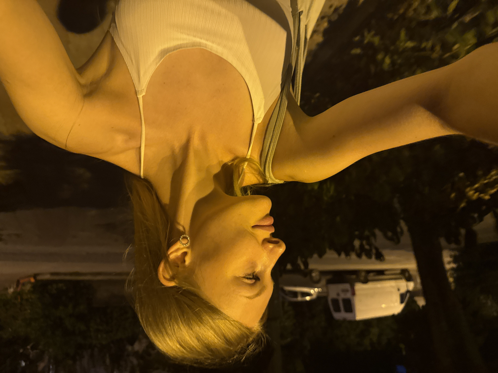 A young woman with blonde hair, wearing a white camisole and earrings, is pictured from the chest up, in a profile view facing left. She is standing outdoors at night, with a street and vehicles in the background. The environment suggests a public area within the city of Kemer, Turkey, likely a street with some greenery and parked vehicles. The lighting indicates artificial streetlights illuminating the scene.
