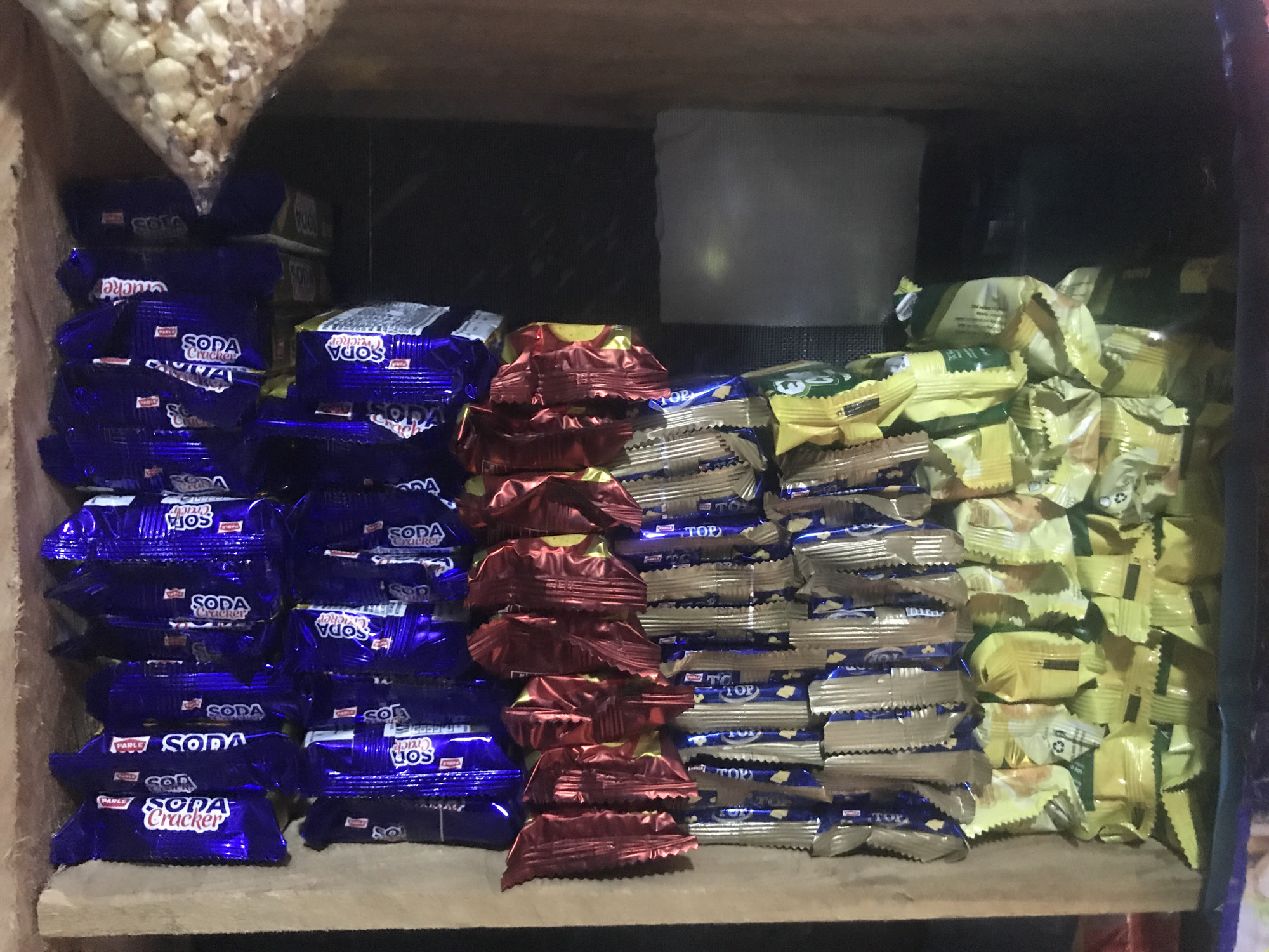 This image shows a shelf in a shop, likely a convenience store or a small supermarket, in New Stadium, Nigeria. The shelf is stocked with various packaged snacks, primarily biscuits and crackers. In the foreground, a stack of dark blue packages labeled "SODA Cracker" is visible. To the right of these, there are stacks of red and yellow foil-wrapped snacks, and further to the right, more yellow and green packaged snacks. In the background, there are also some white sheets or packaging. A portion of a bag of popcorn is visible at the top left of the image. The lighting appears to be artificial and somewhat dim. The overall scene depicts a retail environment with a variety of confectionery items displayed for sale.