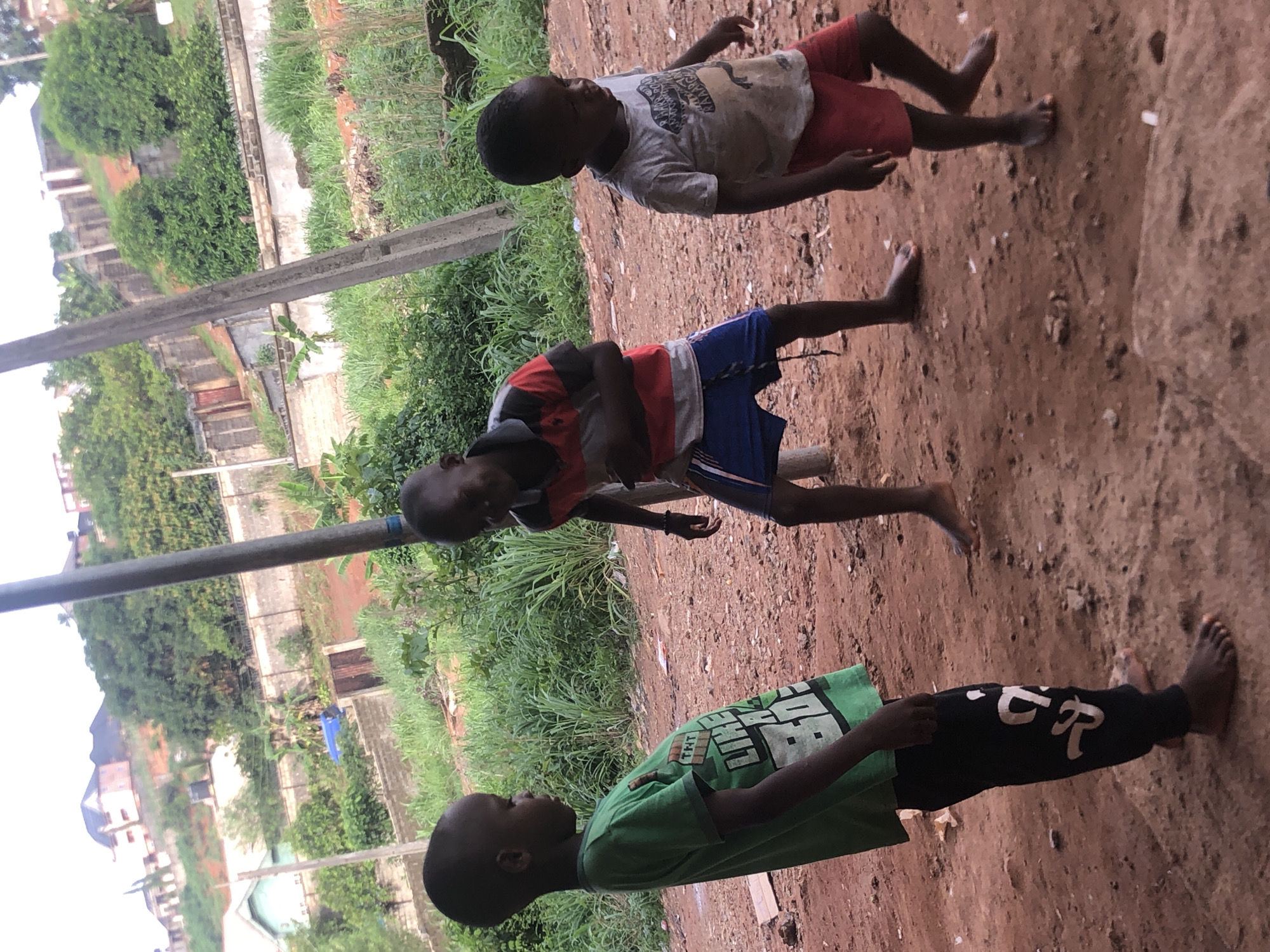 Three young boys play barefoot on a dirt surface outdoors in Nkwelle Ezunaka, Nigeria. One boy, wearing a green T-shirt and black pants with white lettering, is closest to the foreground. Another boy wears a red and gray shirt with blue shorts. The third boy is in a white T-shirt and red shorts. Green grass and vegetation are visible in the background. A metal pole and buildings are also in the background.