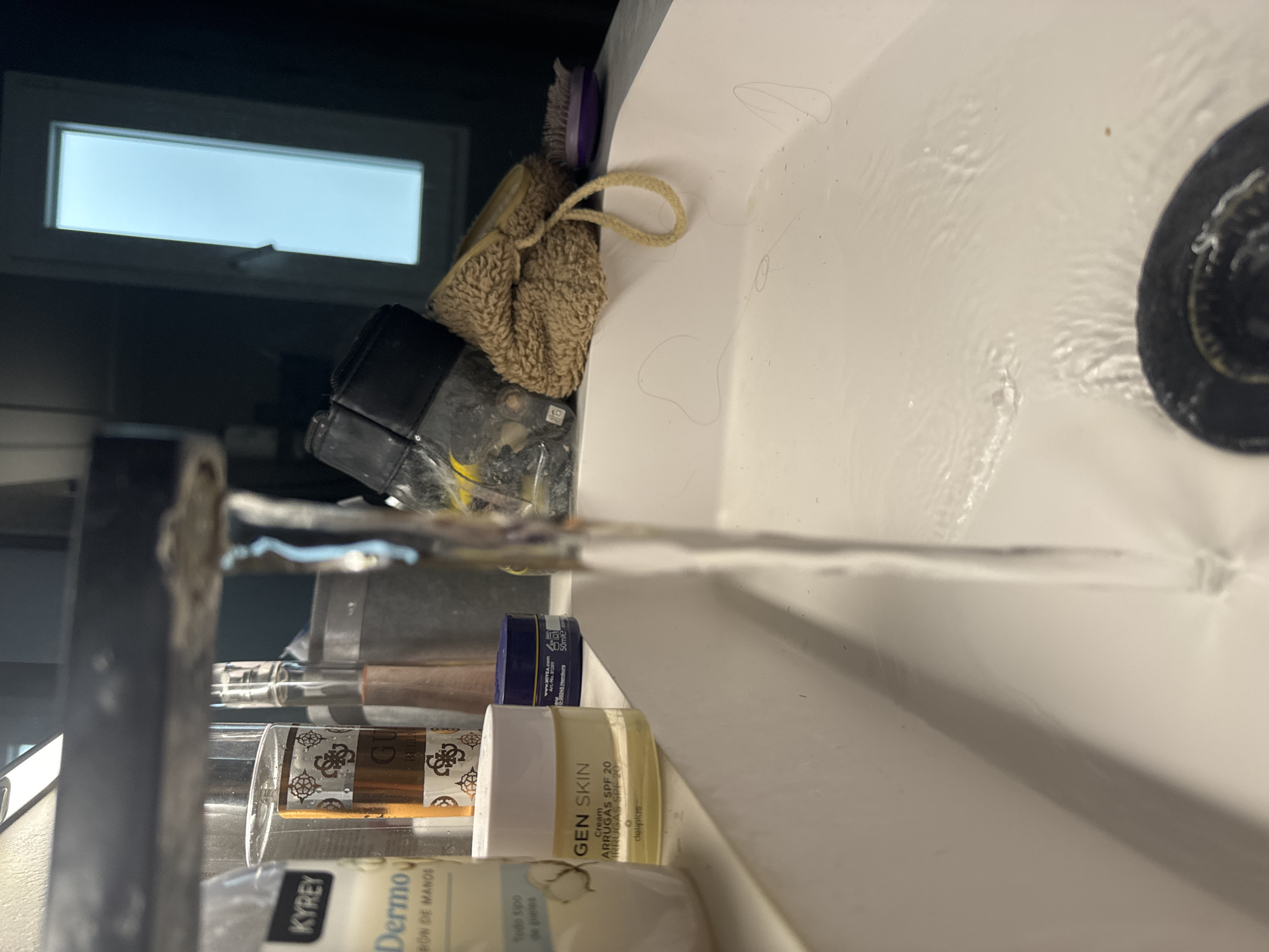 The image depicts a close-up view of a sink in what appears to be a bathroom. Water is running from a faucet, creating ripples on the white ceramic surface of the sink. Several toiletry items are visible on the edge of the sink, including a bottle labeled "KYREY Dermo," a bottle with a "GUESS" logo, and a small jar of "GEN SKIN" cream. In the background, a window with a bright light is visible, and to the right, there's a fuzzy brown object with a string, possibly a loofah or washcloth, and a small purple brush. The overall impression is of a functional bathroom sink area, likely in a residential setting, possibly in the city of Murcia, Spain, given the context provided, though the image itself does not contain explicit geographic identifiers.