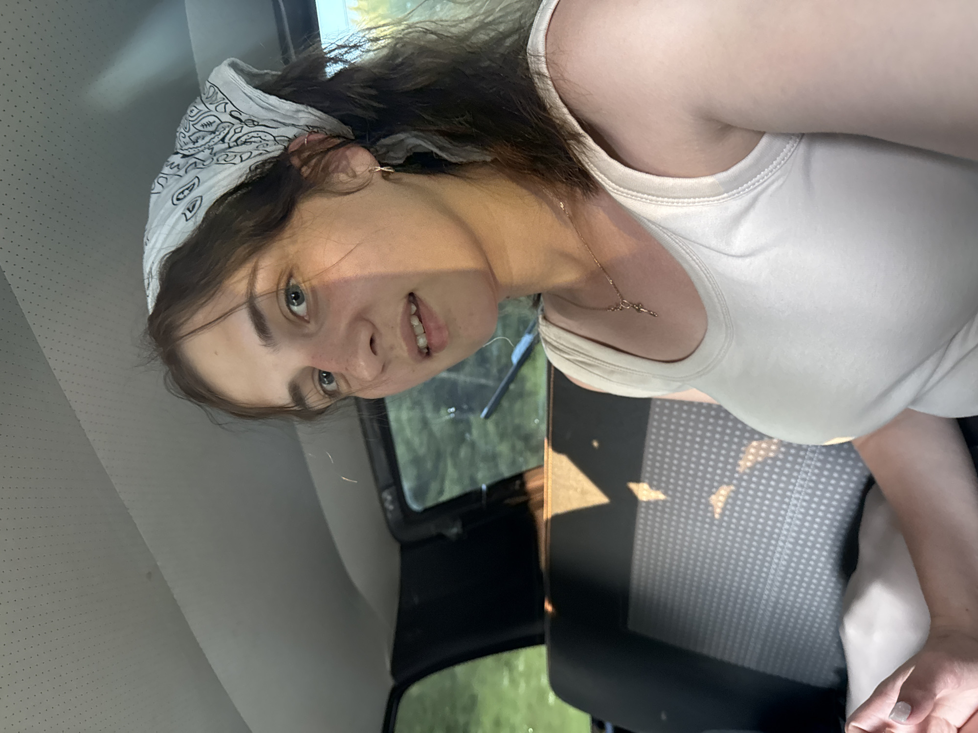 A young Caucasian woman with brown hair, wearing a white tank top and a white bandana, is seated in the backseat of a car. She is looking towards the camera with a slight smile. Sunlight is casting shadows on her face and the car's interior. The background shows the interior of a car, including the backseat and windows, with greenery visible outside. The image appears to be a selfie taken inside the vehicle.