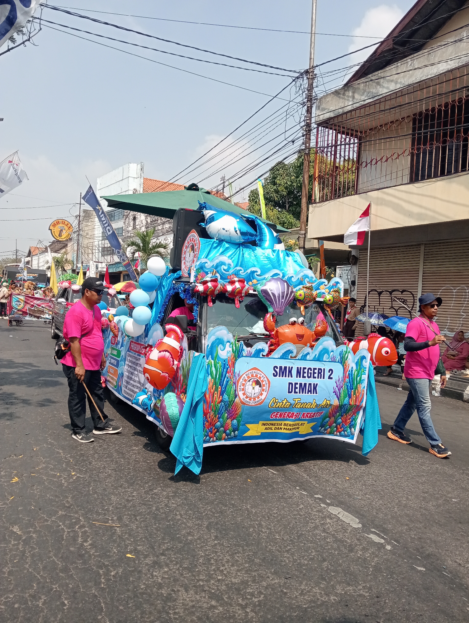 The image shows a parade float decorated with an underwater theme, featuring balloons shaped like fish, a crab, and coral. The float is based on a vehicle and is adorned with blue fabric to represent water. A banner on the float reads "SMK NEGERI 2 DEMAK CINTA TANAH AIR GENERASI KREATIF INDONESIA BERDAULAT ADIL DAN MAKMUR." Two men in pink shirts are near the float, one holding a stick. One man is walking to the right of the float, and another is standing on the left. Several other people are visible in the background, some watching the parade and others on other floats. The scene appears to be taking place on a street in the city of Bintoro, Indonesia, with buildings and utility poles lining the road. The sky is clear and the sun is shining, suggesting it is daytime.
