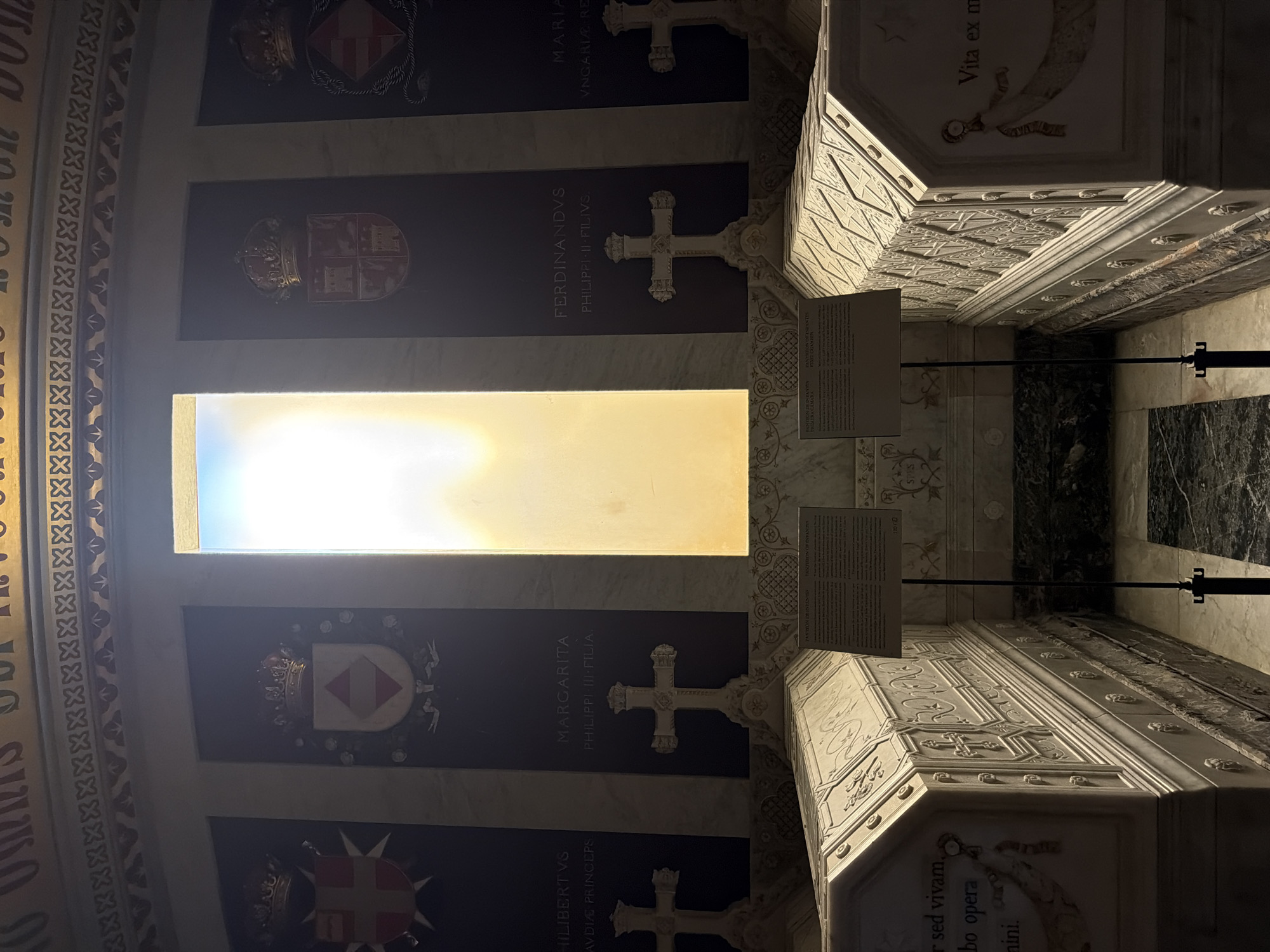 The image depicts the Royal Pantheon of the Monastery of San Lorenzo de El Escorial, located in San Lorenzo de El Escorial, Spain. The scene is dimly lit, with a bright rectangular window at the back of the chamber casting a strong backlight. In the foreground, two ornate sarcophagi are visible, made of white marble and decorated with intricate carvings. The sarcophagus on the left has text inscribed on its side, reading "er sed vivam, obo opera minini." The sarcophagus on the right has text that reads "Vita ex m".
Behind the sarcophagi, on the walls, are four dark panels, each featuring a royal coat of arms and a crown at the top. The names "PHILIBERTVS", "MARGARITA", "FERDINANDVS", and "MARIA" are inscribed on these panels, along with their familial relationships. Between these panels are two large white crosses, also carved from marble, mounted on ornate bases. The floor appears to be made of polished dark stone with lighter marble patterns. Two metal stands with informational plaques are positioned between the sarcophagi. The overall atmosphere is solemn and monumental, characteristic of a royal burial site.