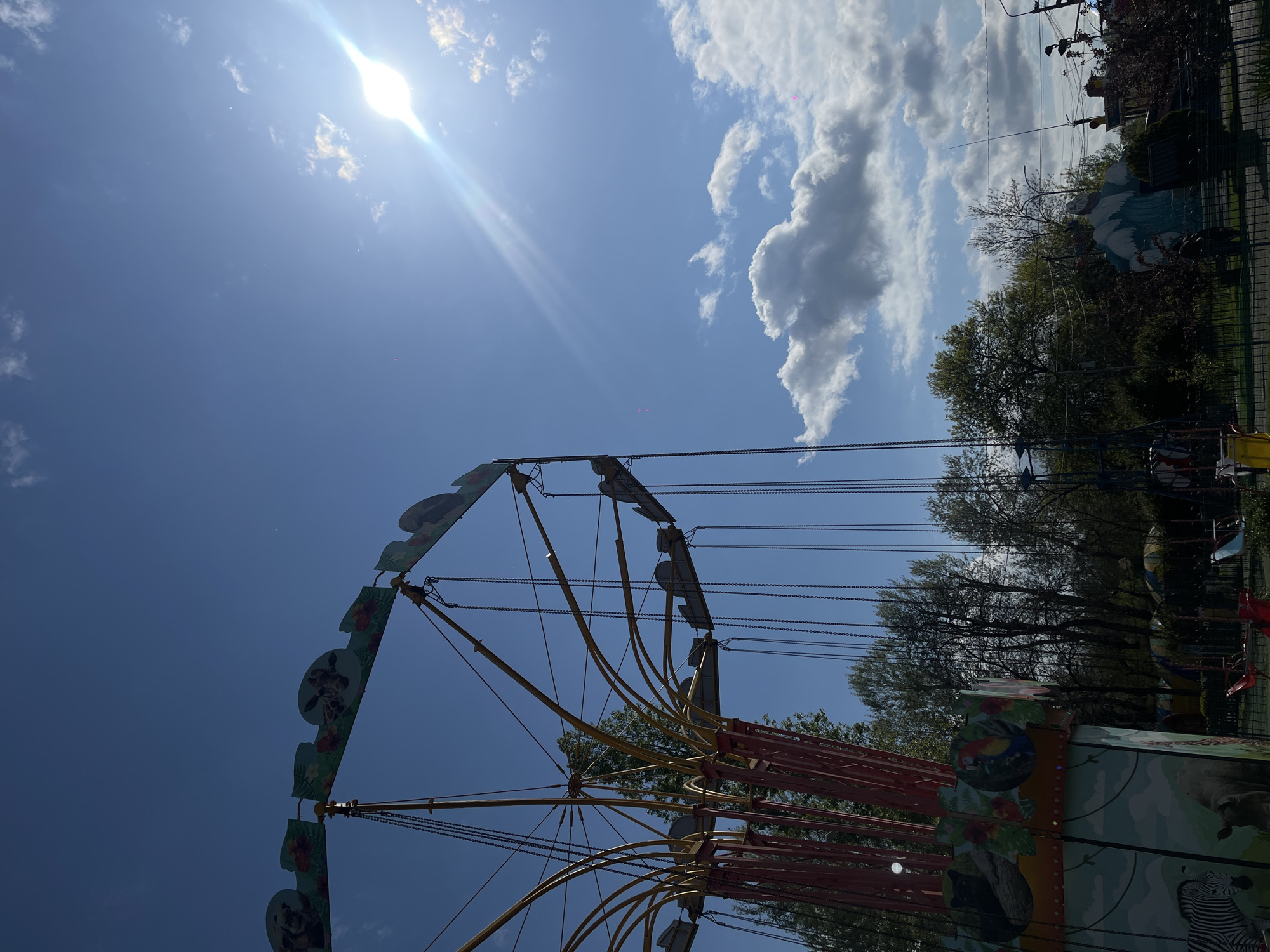 A view of a sunny sky with some clouds, partially obscured by a swing carousel in an amusement park. The carousel features animal-themed decorative panels. In the background, trees and park foliage are visible, and faint structures and infrastructure can be made out at the edge of the park. The photograph seems to have been taken in Krasnodar, Russia.