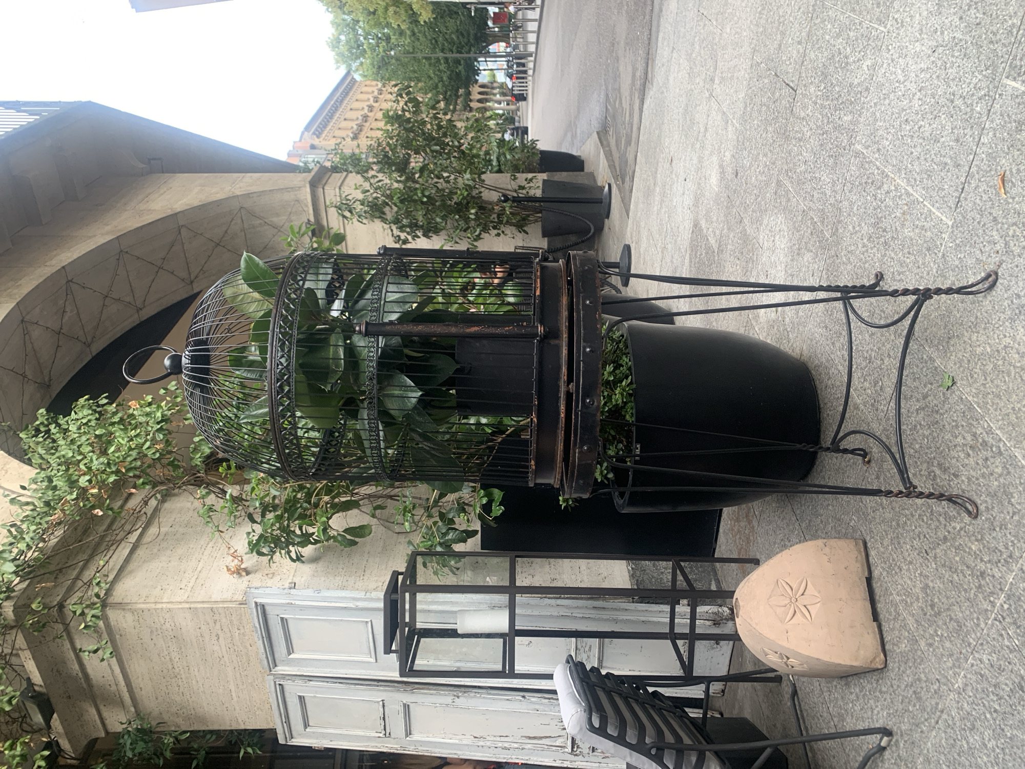 This is a medium shot of an outdoor cafe or seating area, likely in Lugano, Switzerland, judging by the architectural style and general ambiance. The foreground is dominated by a large, ornate black metal birdcage on a stand, containing a lush green plant. Next to the birdcage is a tall, thin black metal lantern with a white candle inside, and further left, a portion of an outdoor chair with a grey cushion is visible. Behind these objects, a cream-colored textured wall with an arched overhang and some weathered white doors can be seen. To the right, a street with parked cars and pedestrians stretches into the background, bordered by buildings and trees. The ground is paved with large, light-colored square tiles. The overall scene suggests a tranquil, slightly rustic urban setting.