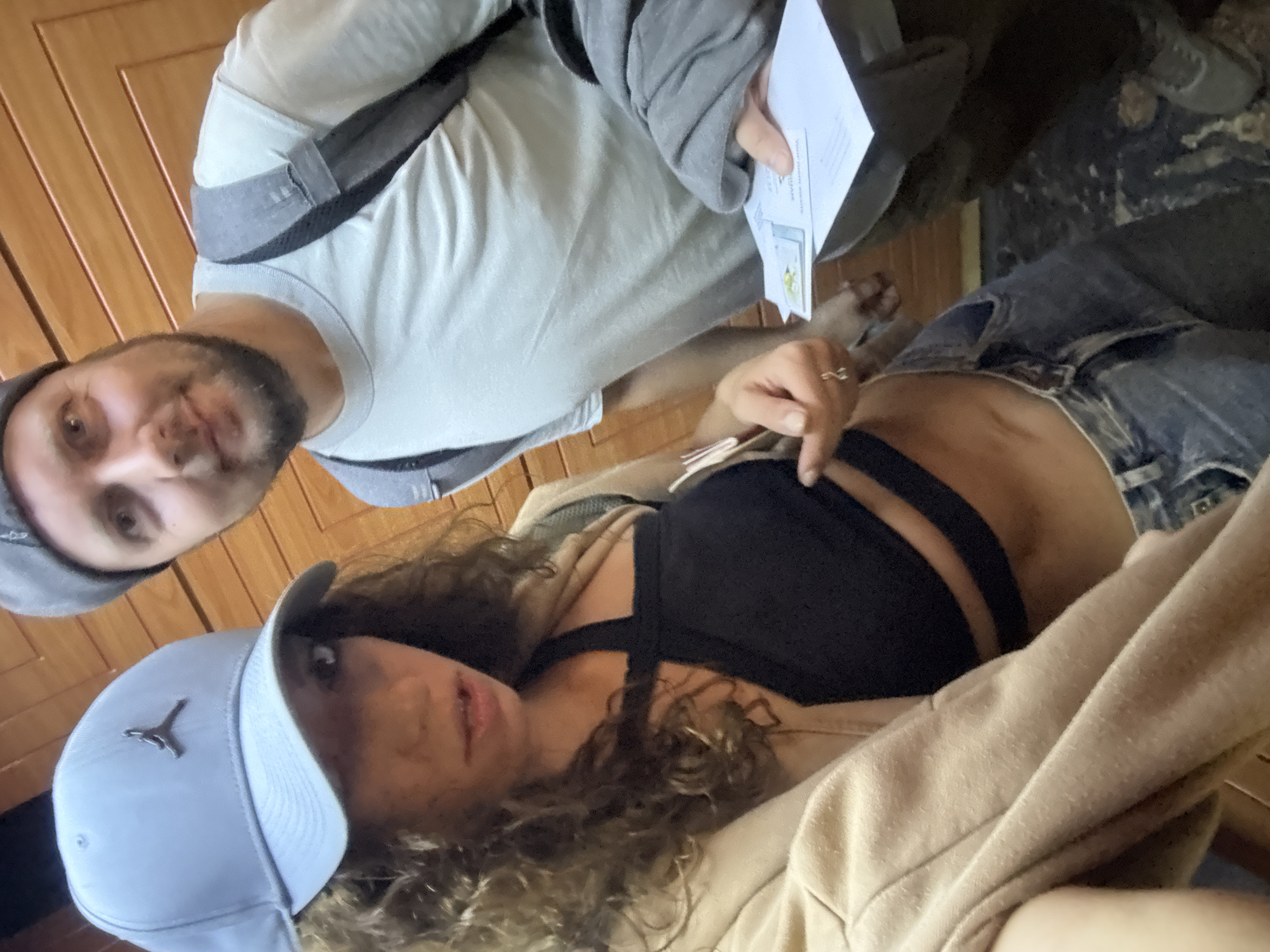 A selfie captured from a low angle features two individuals. A man with a trimmed beard and a grey cap is visible in the upper-left, wearing a light grey t-shirt and a backpack. He is holding a white card in his right hand. Adjacent to him is a woman with curly hair, wearing a light blue Jordan cap and a black bra top. She is partially covered by a beige jacket or robe. Her left hand is visible near her midriff. The background consists of wood paneling and a dark, textured surface. The location is purportedly in Đà Nẵng, Vietnam.