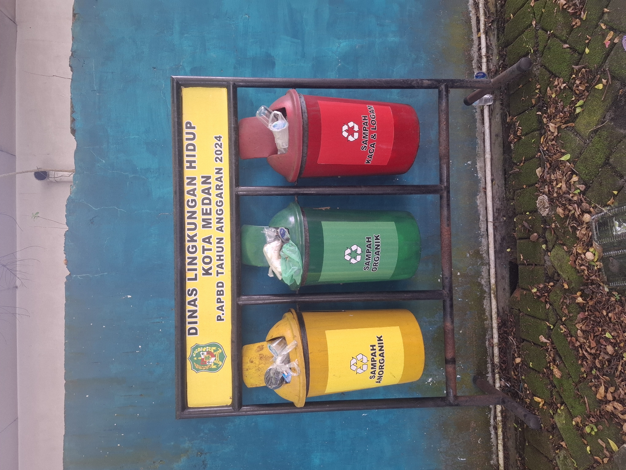 The image shows a trash receptacle stand with three bins: one yellow, one green, and one red, against a weathered blue wall. The yellow bin is labeled "SAMPAH ANORGANIK" (Inorganic Waste), the green bin is labeled "SAMPAH ORGANIK" (Organic Waste), and the red bin is labeled "SAMPAH KACA & LOGAM" (Glass & Metal Waste). Above the bins is a yellow sign with the text "DINAS LINGKUNGAN HIDUP KOTA MEDAN P. APBD TAHUN ANGGARAN 2024" which translates to "Environmental Agency of Medan City P. APBD Fiscal Year 2024". The ground in front of the bins is covered with moss, leaves, and debris. The overall scene suggests an outdoor public area in Medan, Indonesia, likely a street or park, with a focus on waste segregation. There are no people visible in the image.