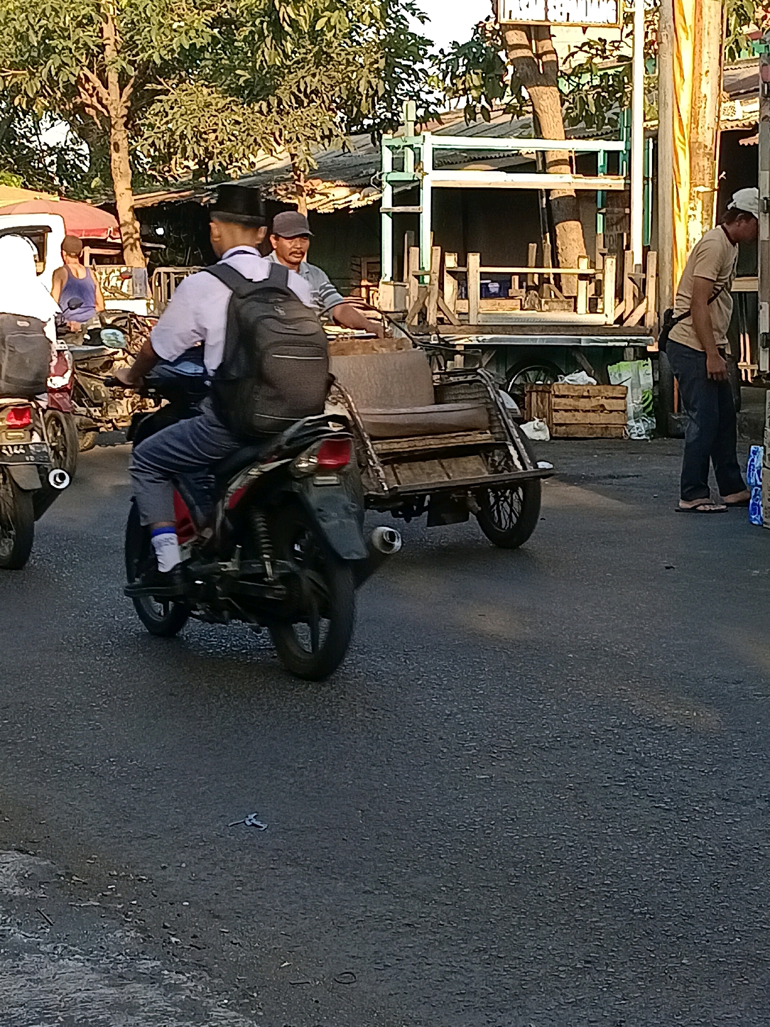 In Wonocolo, Indonesia, on a paved road, a man wearing a white shirt, blue pants, and a black hat rides a motorcycle. He has a black backpack on. The motorcycle is red and black. Behind him, a rickshaw is attached to another motorcycle. The rickshaw has a brown seat and a black wheel. To the right of the rickshaw, a man in a tan shirt and blue jeans stands next to a white truck. The truck has wooden crates on it. The environment is outdoors with trees and buildings in the background. The sun is shining.