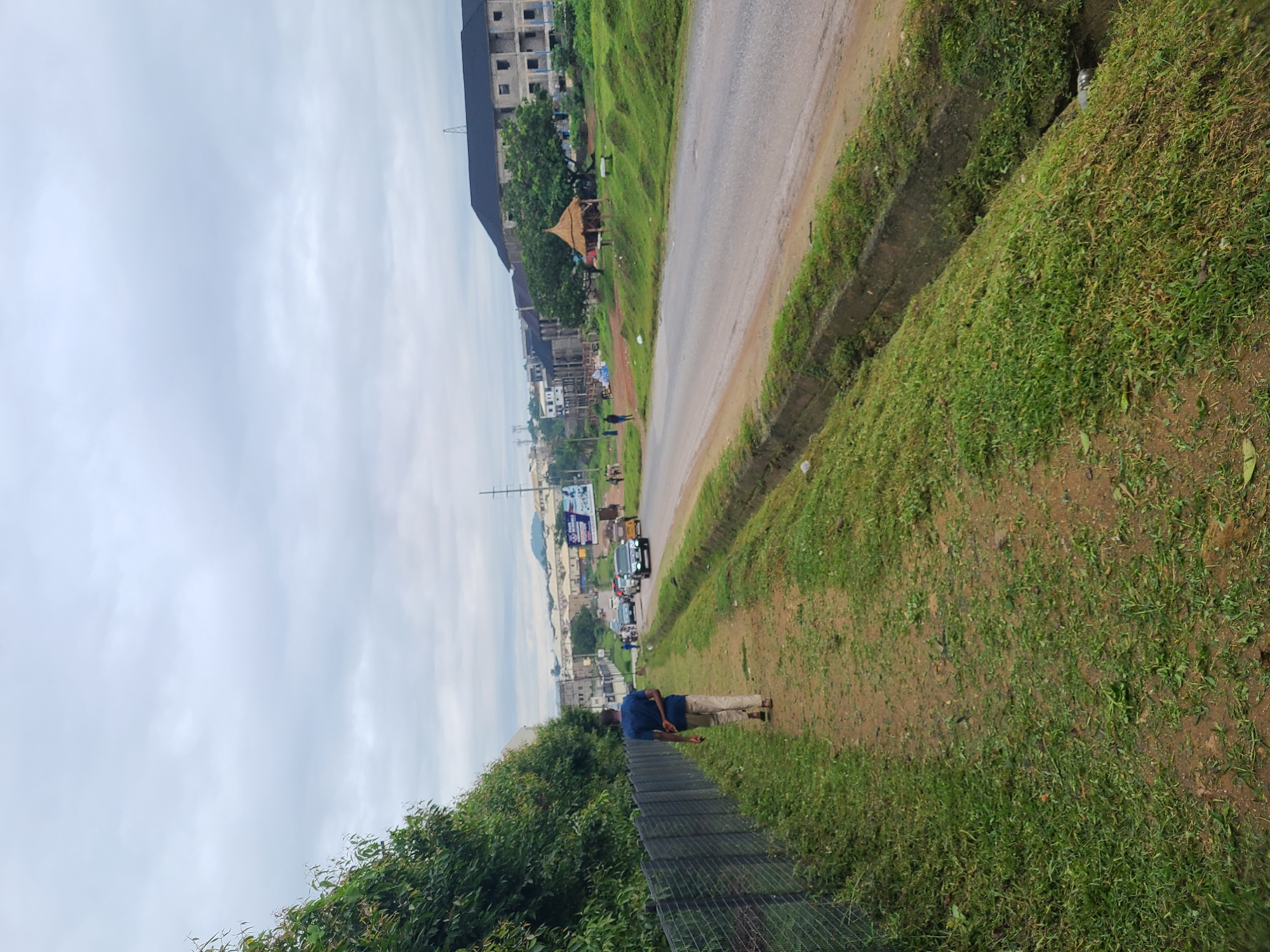 A medium-shot captures a person walking away from the camera on a dirt path lined with a metal fence and lush green vegetation on the left. The path runs parallel to a paved road on the right, which has a drainage ditch. The road is moderately busy with several vehicles, including cars, a van, and a tricycle. In the background, there are multi-story buildings, some appearing to be under construction, and a small thatched hut. The landscape extends to a gently rolling hill under an overcast sky. The scene appears to be in Abuja, Nigeria, given the context of similar streetscapes and architecture. The overall impression is an urban fringe setting, possibly a developing area.