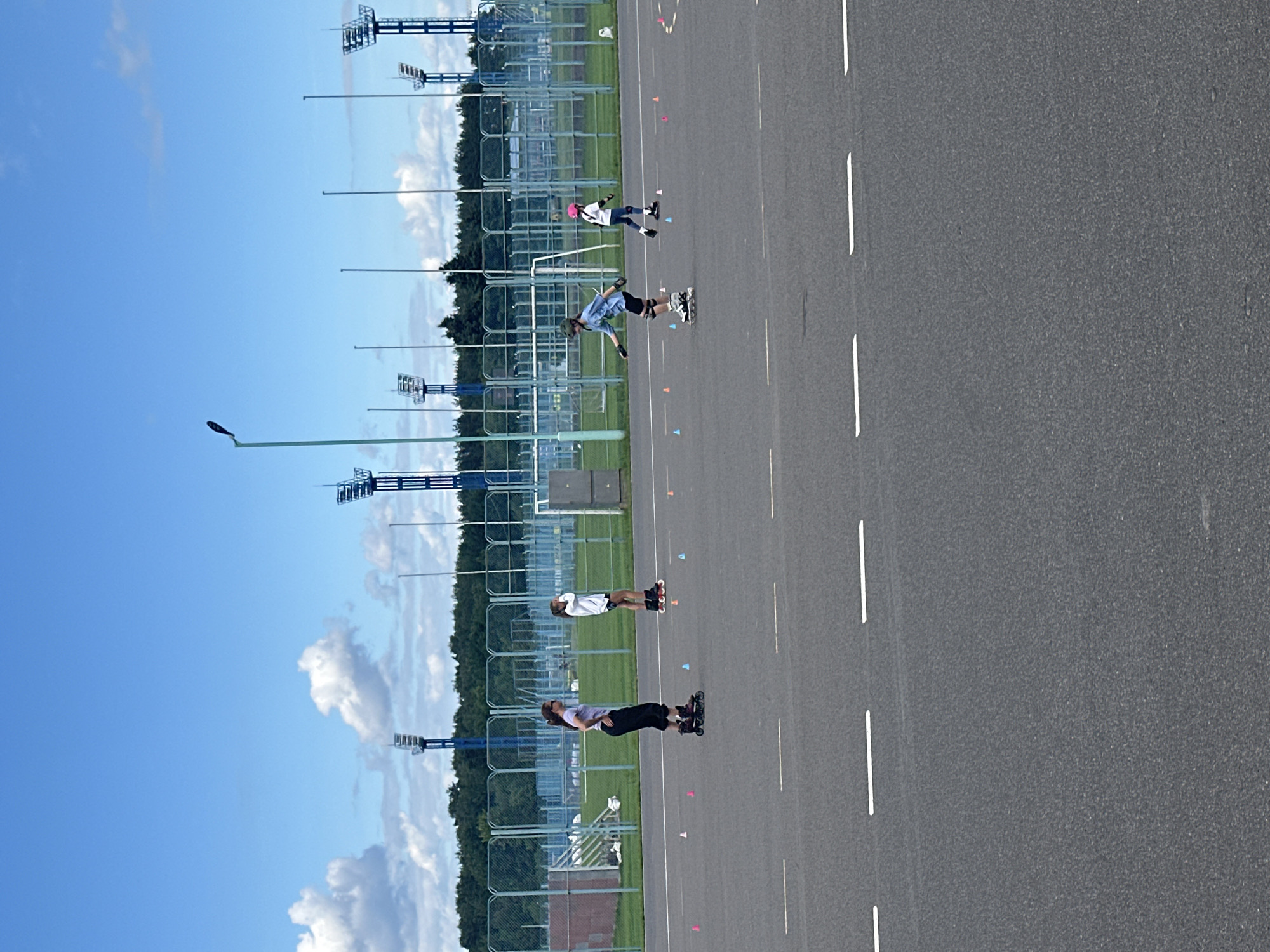 A group of people are inline skating on an asphalt road. The image is oriented vertically, with the road taking up the right two-thirds of the frame and the sky and distant landscape filling the left third. There are several people on roller skates, some wearing protective gear such as helmets. In the foreground, two individuals are skating, one in a white shirt and black shorts, and another in a dark long-sleeved shirt and black pants. Further back, another person is skating in a blue shirt and shorts. In the distance, there are what appear to be sports fields with fencing and tall light poles. The sky is bright blue with scattered white clouds, suggesting it is daytime and the weather is fair. The asphalt road has white lane markings. The overall scene appears to be a recreational activity taking place outdoors in a somewhat urban or sports complex environment. There is no visible text in the image.