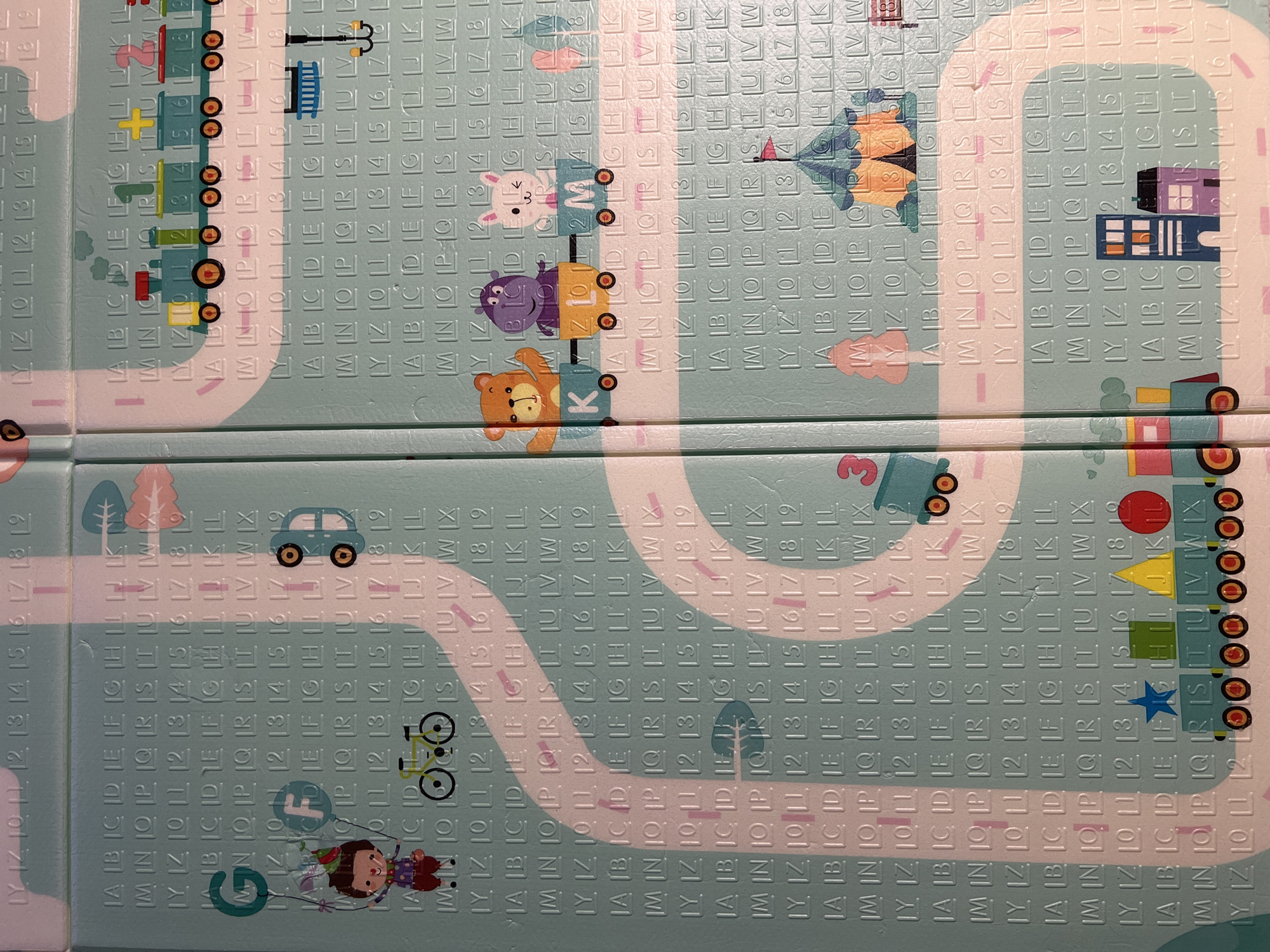 The image shows a close-up of a colorful children's play mat with various cartoon-like illustrations and educational elements. The mat is made of interlocking foam tiles.
Illustrations include:
* A train with animal passengers (bear, hippo, rabbit)
* A blue car
* A child with a balloon
* A bicycle
* A house
* A circus tent
* Numbered and lettered pathways
The mat also features repeated rows of letters (A-Z) and numbers (1-19). The tiles are light blue with white pathways and various colorful graphics. The overall impression is that of a play area designed to be both entertaining and educational for young children.