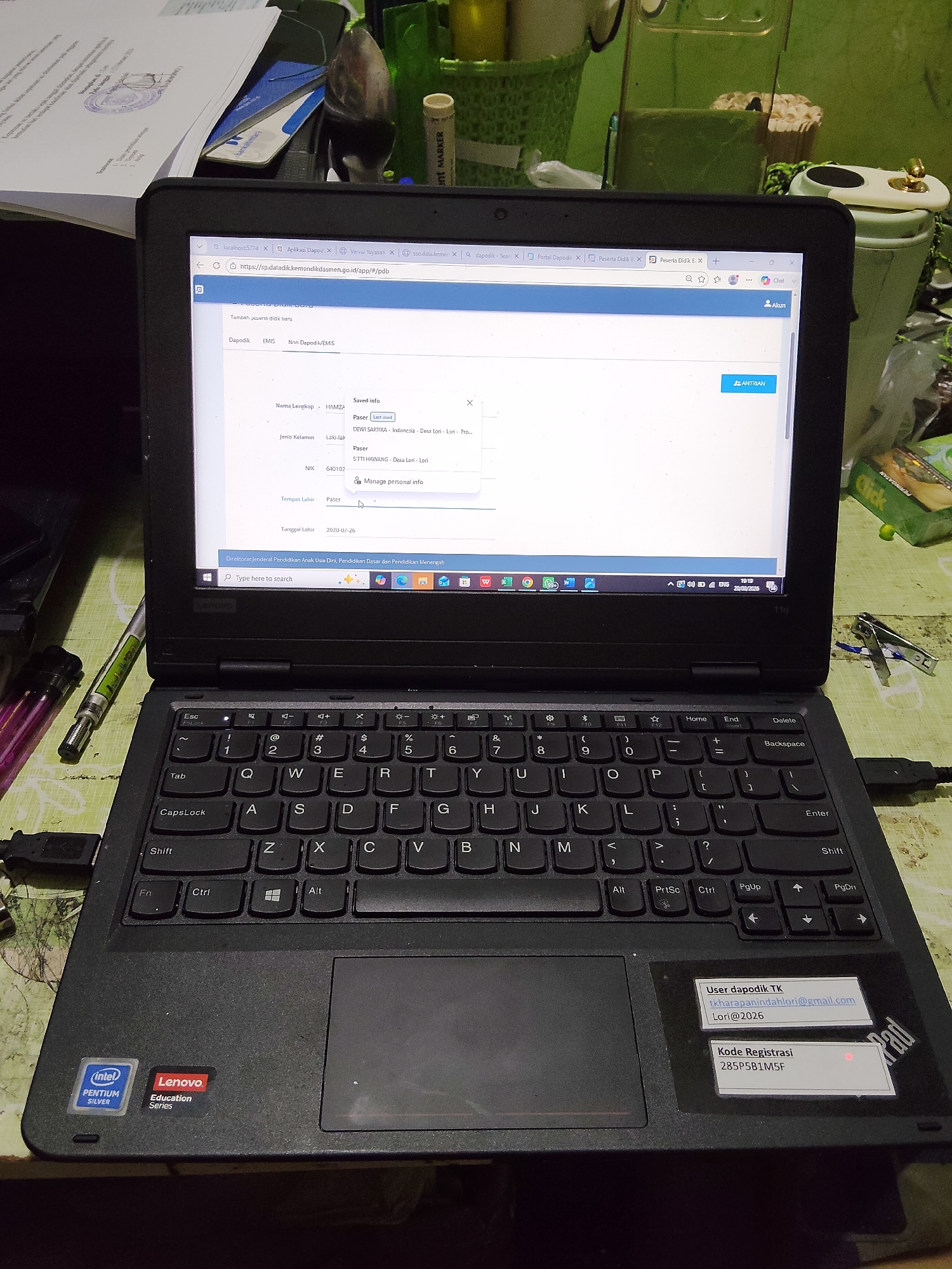 The image shows a laptop computer open and displaying a website titled "Aplikasi Dapodik". The website appears to be a form for entering student information, with fields for name, gender, NIK, and date of birth. The laptop is on a table, with other objects visible in the background, including papers, a stapler, and various containers. A sticker on the laptop shows the "Intel Pentium Silver" and "Lenovo Education Series" logos. Another sticker on the palm rest of the laptop displays "User dapodik TK" with an email address and username, and "Kode Registrasi" with a registration code. The overall environment appears to be an indoor setting, possibly a home office or classroom, with natural light entering from an unseen window. The city of Lori, Indonesia, is not directly indicated in the image, but the context of the Dapodik application, which is used in Indonesian educational institutions, suggests the location might be within Indonesia.