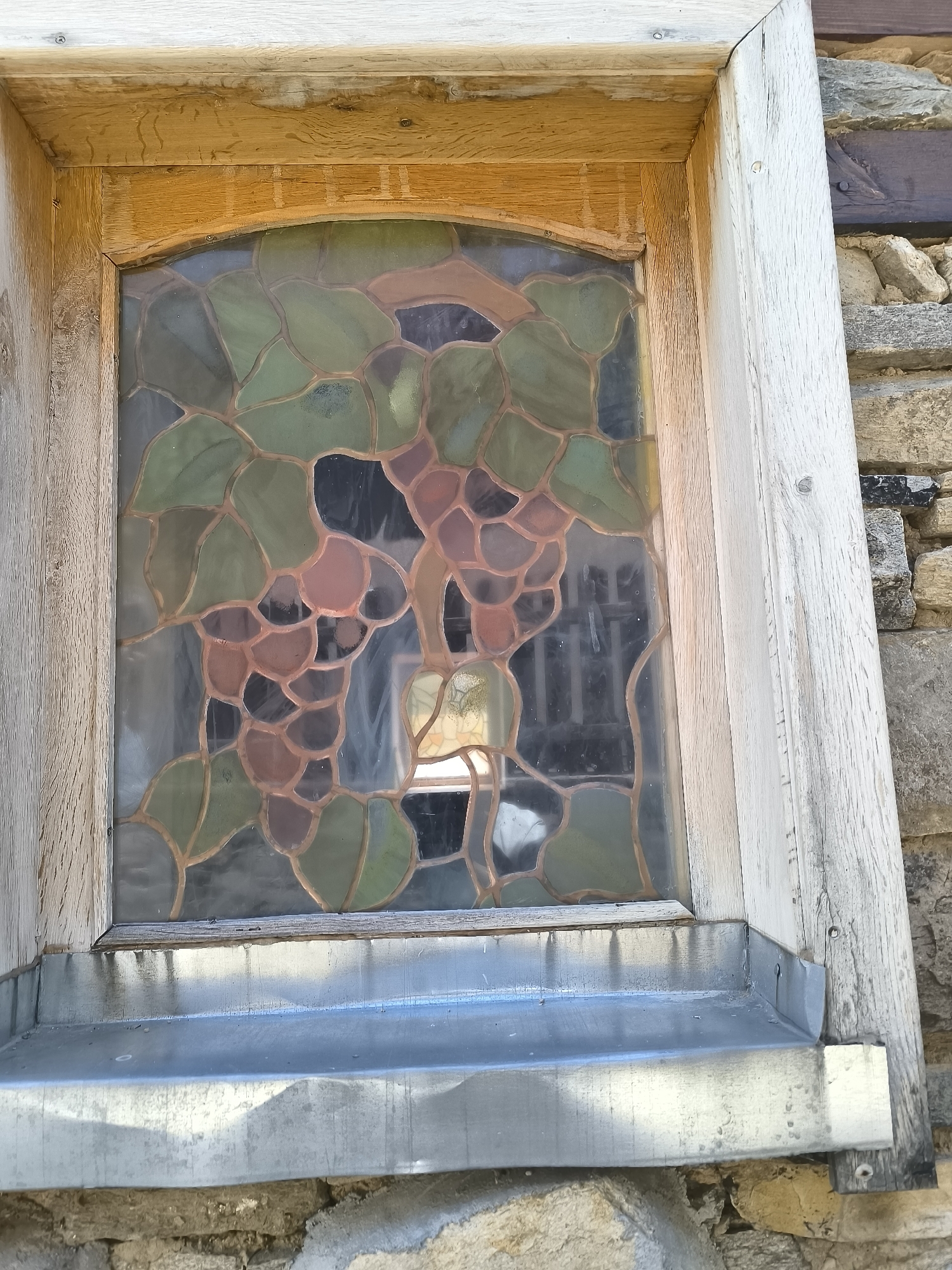 The image shows a close-up of a small stained-glass window depicting grapes and leaves, framed by weathered wooden beams. The window is set into a stone wall. A metal ledge runs along the bottom of the window. The style of the stained glass and the architectural elements suggest a traditional building, likely a house or church. The location is identified as Leshten, Bulgaria. The image is taken from a slightly low angle, looking up at the window.