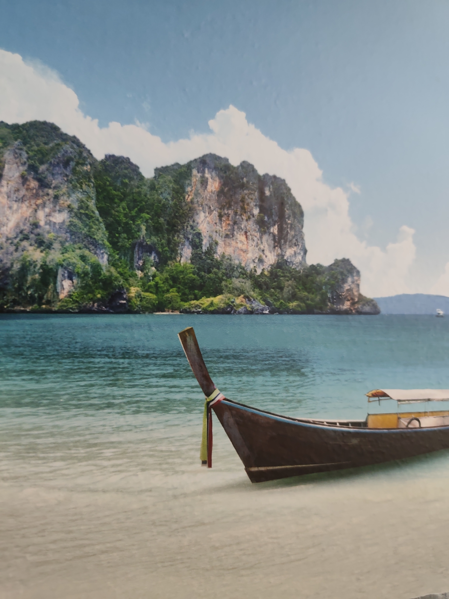 The image captures a serene tropical coastal landscape, not located within a city. In the foreground, a traditional dark-hulled longtail boat, featuring a light blue trim and a yellow and white canopy section, rests partially on a light, wet sandy beach and extends into clear, shallow, turquoise water. A decorative multi-colored fabric streamer (red, yellow, and green) is tied to the tall, upright stern rudder or engine mount. The water transitions from shallow, rippling clear turquoise near the shore to a deeper blue-green further out. The midground is dominated by several large, steep limestone karsts (cliffs) densely covered in lush green tropical vegetation, rising dramatically from the sea. In the far distance on the right side of the frame, a small white boat is visible on the water. The sky above is a bright light blue, punctuated by scattered fluffy white cumulus clouds. No people are visible in the scene.
