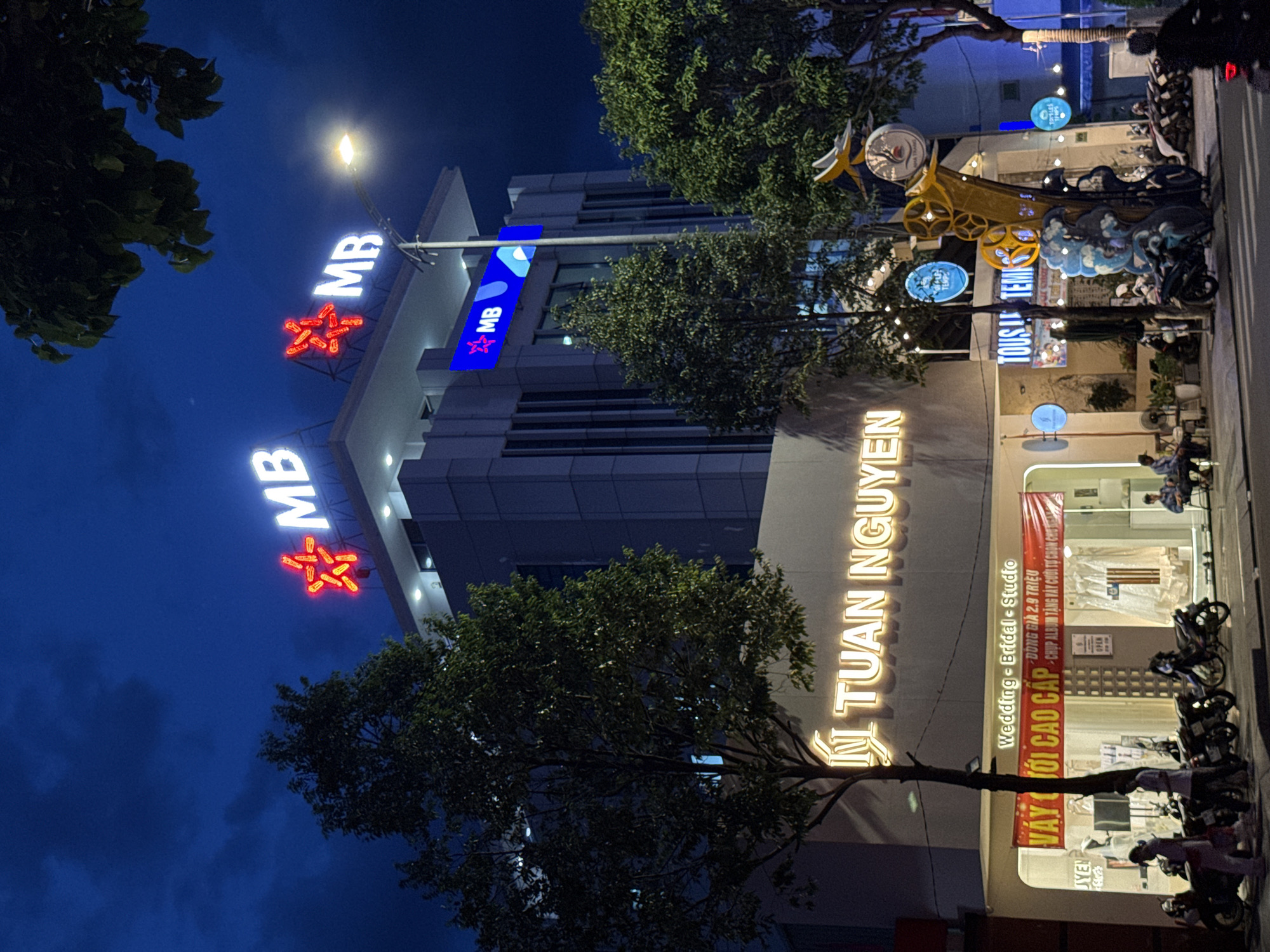 This image captures a nighttime street scene in Nha Trang, Vietnam. Dominating the upper portion of the frame is a modern, multi-story building with prominent "MB" signage illuminated with red stars on its rooftop. Further down, a large, glowing sign reads "TUAN NGUYEN," likely indicating a business. Below this, a shop with a wide storefront displays wedding and bridal studio services, with a red banner proclaiming "VÁY CƯỚI CAO CẤP" (High-end wedding dresses). Inside the studio, wedding dresses are visible. The street in front of the shops is lined with parked motorcycles. Several people are present, some walking along the sidewalk, others sitting outside, and one person is riding a motorcycle on the street. Trees with lush green foliage are interspersed throughout the scene, and decorative lighting is visible. The sky is dark blue, indicating it is nighttime. A streetlamp is illuminated, casting light on the scene.