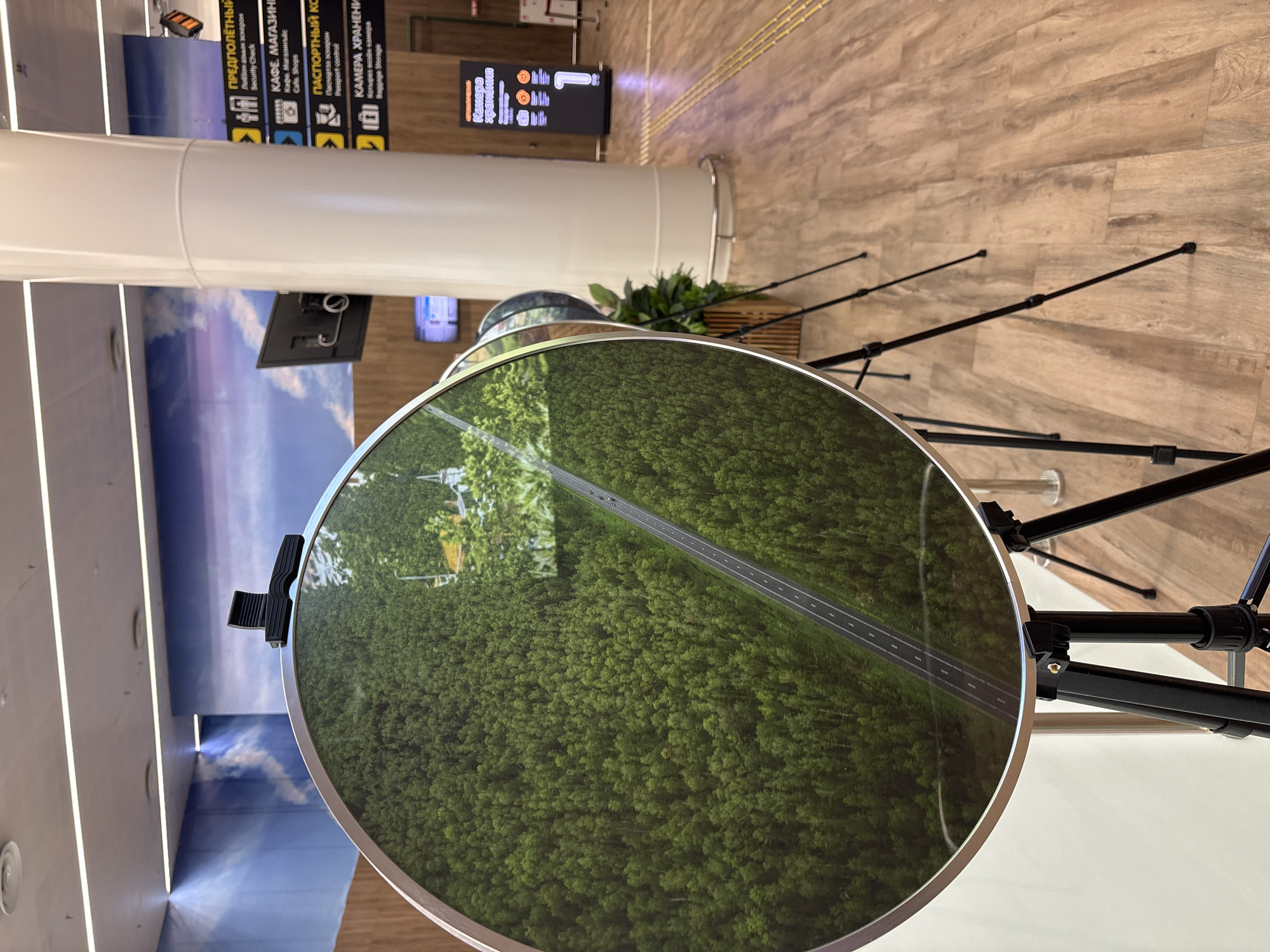 This media file captures an interior scene, likely an airport terminal or a public transit station, given the directional signage. The main subject is a large, circular reflective surface that displays a vibrant aerial view of a dense green forest bisected by a grey asphalt road. The reflection suggests this is a display or a decorative element.
In the background, typical airport elements are visible. To the left, a large white pillar dominates the frame, with a digital display or mural depicting a sky with clouds on the wall behind it. Above and to the left of the circular display, directional signs in Cyrillic script are mounted on a wall, indicating "ПРЕДПОЛЁТНЫЙ" (Pre-flight), "КАФЕ. МАГАЗИНЕ" (Cafe. Shops), "Пастерная аскарен" (Passport control), and "КАМЕРА ХРАНЕНИ" (Baggage Storage). These signs, along with the modern lighting and polished floor, confirm the setting is a transportation hub.
The circular display is supported by a black tripod structure, implying it might be a temporary or portable exhibit. A small black clip is attached to the top edge of the circular display. A potted green plant is visible behind the display, adding a touch of nature to the otherwise man-made environment. The lighting appears to be artificial, suggesting an indoor environment, likely during the day. There are no people visible in the frame, emphasizing the objects and the setting. The overall impression is one of modern public space with a visually interesting display.