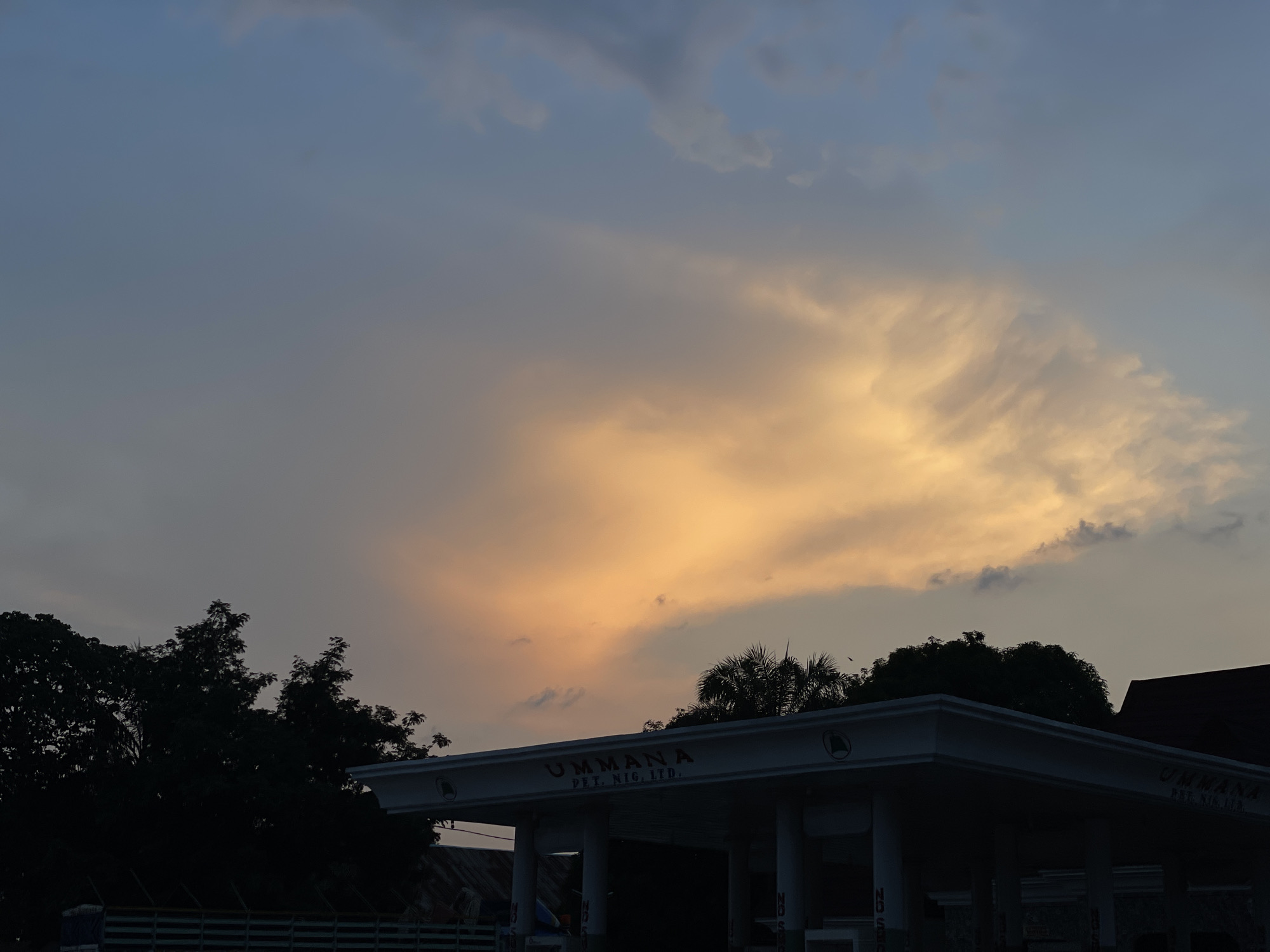 The image depicts a view in Akwanga, Nigeria, at dusk or dawn. The sky is dominated by shades of blue and orange clouds. Below the horizon line are silhouetted trees and the roofline of a building. The building is a gas station branded "UMMANA PET. NIG. LTD." with visible support columns. The scene is dimly lit, suggesting either the early morning or late evening. No people are visible.