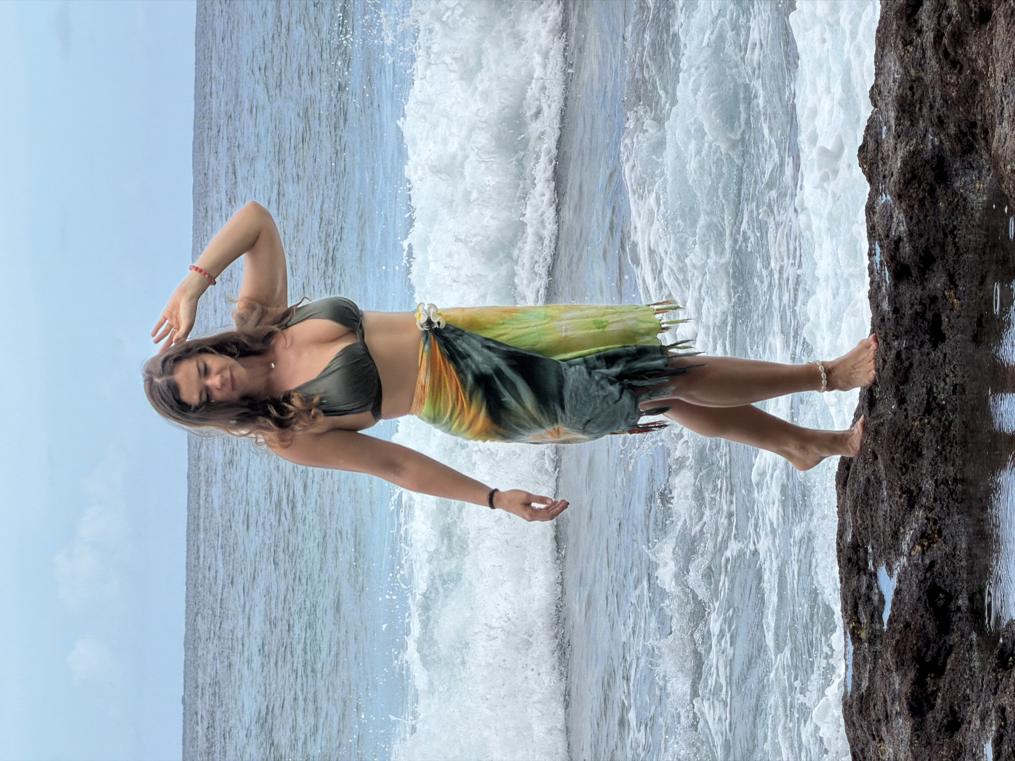 A young woman stands elegantly on a rugged, dark rock formation at the edge of the ocean, likely in Nusa Penida, Indonesia. The image, presented in a full-body shot, captures her looking directly at the viewer with a gentle, serene smile. Her long, dark brown hair cascades over her shoulders.
She is dressed in a dark green bikini top and a flowing sarong-style skirt that features vibrant green, yellow, and orange hues, possibly with a tie-dye pattern. Accessories include a red bracelet on her left wrist, a black bracelet on her right wrist, and a white shell anklet on her left ankle. Her pose is graceful, with her left arm raised, hand near her head, and her right arm extended downwards, palm open.
Behind the woman, the vast ocean unfolds under a light blue or slightly overcast sky, stretching towards the horizon. Dynamic white waves crash and foam, creating an energetic backdrop to the scene. The dark rock formation she stands on occupies the right side of the frame, its textured surface contrasting with the smooth water. The overall atmosphere conveys a sense of peace and connection with nature during what appears to be daytime. No text is visible in the image.
