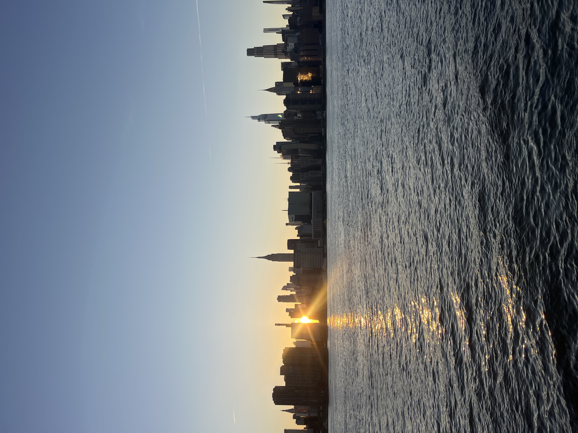 This is a photograph of the Manhattan skyline viewed from across the East River, likely from Brooklyn or Queens, in New York City, USA. The image captures a sunset with the sun positioned behind a cluster of buildings in the mid-ground, casting a bright glare and reflecting intensely on the water's surface. The sky is clear and a pale blue, fading into a lighter shade near the horizon. The water in the foreground is dark with gentle waves, reflecting the golden hues of the setting sun in a shimmering path. The skyline features a variety of skyscrapers, including the iconic Empire State Building, its spire clearly visible. The buildings are silhouetted against the bright sky, with some illuminated by the sun's rays. There are no visible people or boats in the frame. The scene evokes a sense of calm and beauty associated with the end of the day in the bustling city.