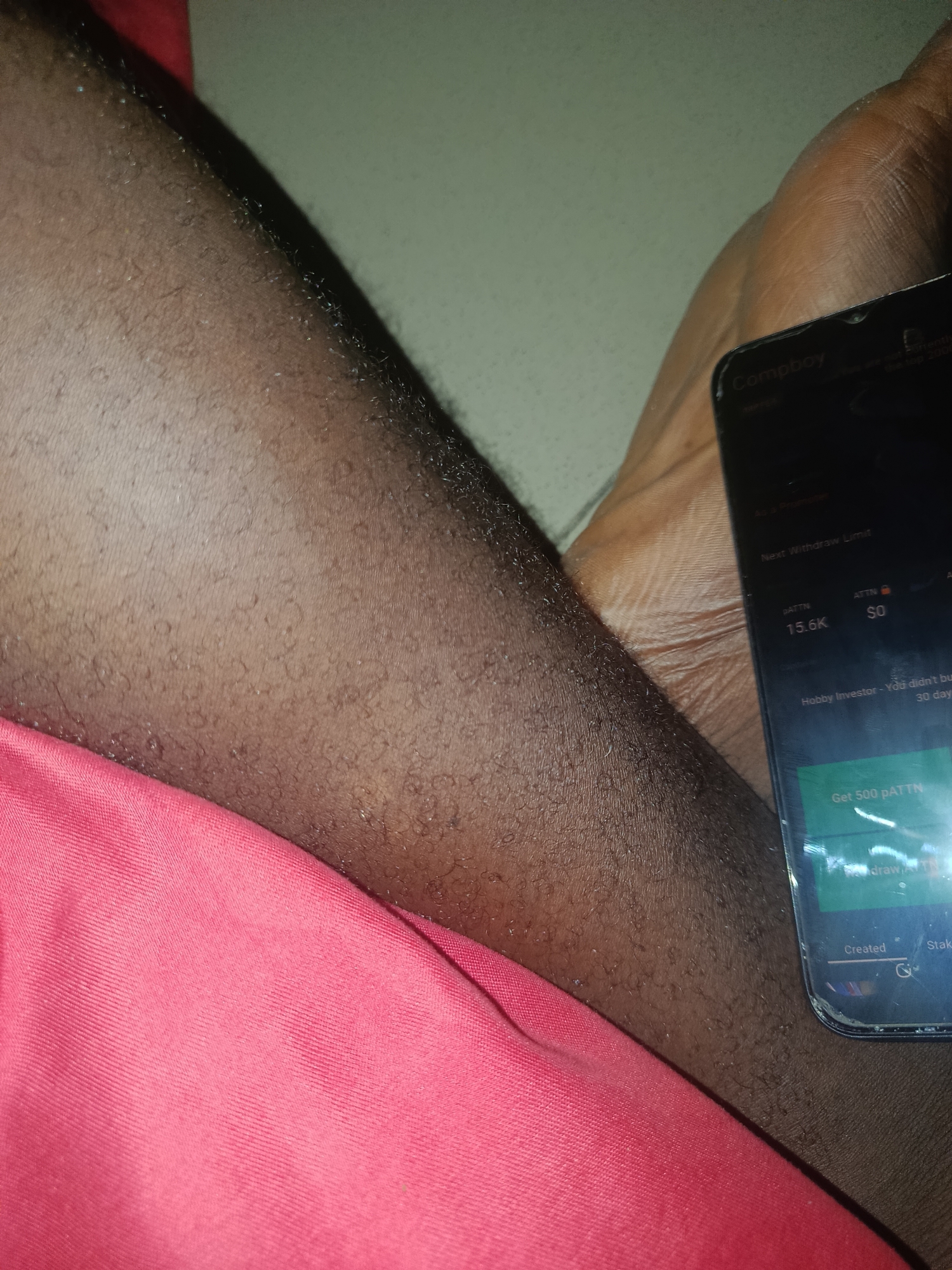 The image displays a close-up of a person's bare leg with dark skin and visible hair follicles, positioned in the foreground. A piece of bright pink fabric, possibly clothing, is visible in the lower left corner, partially obscuring the leg. On the right side, a dark-colored smartphone is held, with its illuminated screen facing the viewer. The phone's screen displays an application interface with text, including "Compboy" at the top, "Next Withdraw Limit," "pATTN 15.6K," "ATTN $0," and "Hobby Investor - You didn't buy... 30 days." A green button prompts "Get 500 pATTN." The phone's screen shows signs of wear, including scratches or cracks near the bottom right. Another body part, likely a foot, is partially visible on the right, holding or resting near the phone. The background consists of a plain, light-colored, out-of-focus surface. The scene is illuminated by artificial light. The location is Unknown, Unknown.