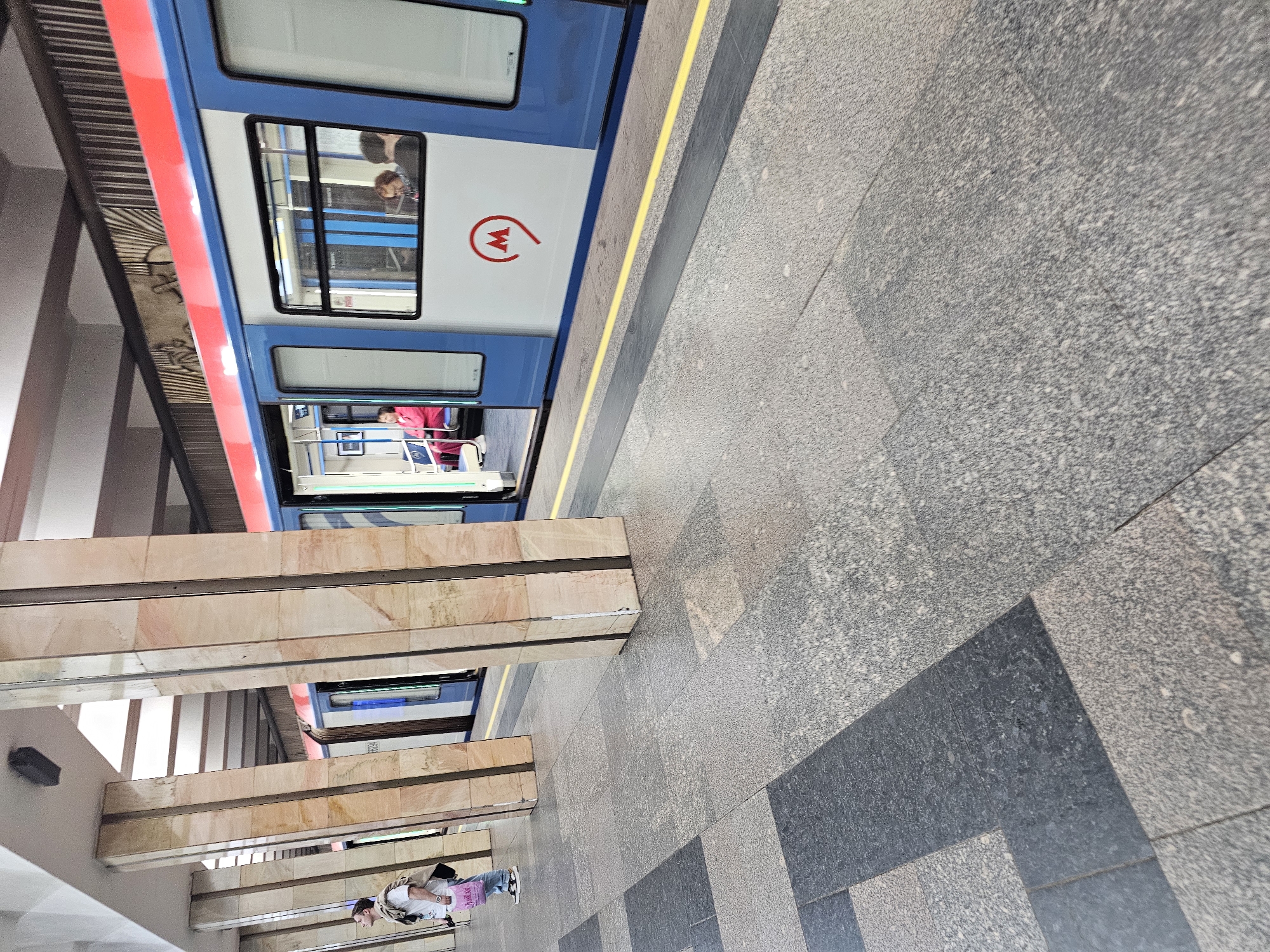 An exterior medium shot captures a metro station platform in an urban environment. The platform is paved with large, square granite tiles in varying shades of gray and black, with a yellow safety line running along the edge. Several large, square columns with polished beige and pink marble cladding support an overhead structure. A metro train with blue and white sides and a red stripe along the top is stopped at the platform, with its doors open. Inside the train, several passengers are visible, including a woman in a red jacket sitting, and a man and woman in the background. On the platform, a person wearing a white t-shirt and jeans stands near the columns, holding a pink shopping bag. The scene is brightly lit, suggesting it is daytime.