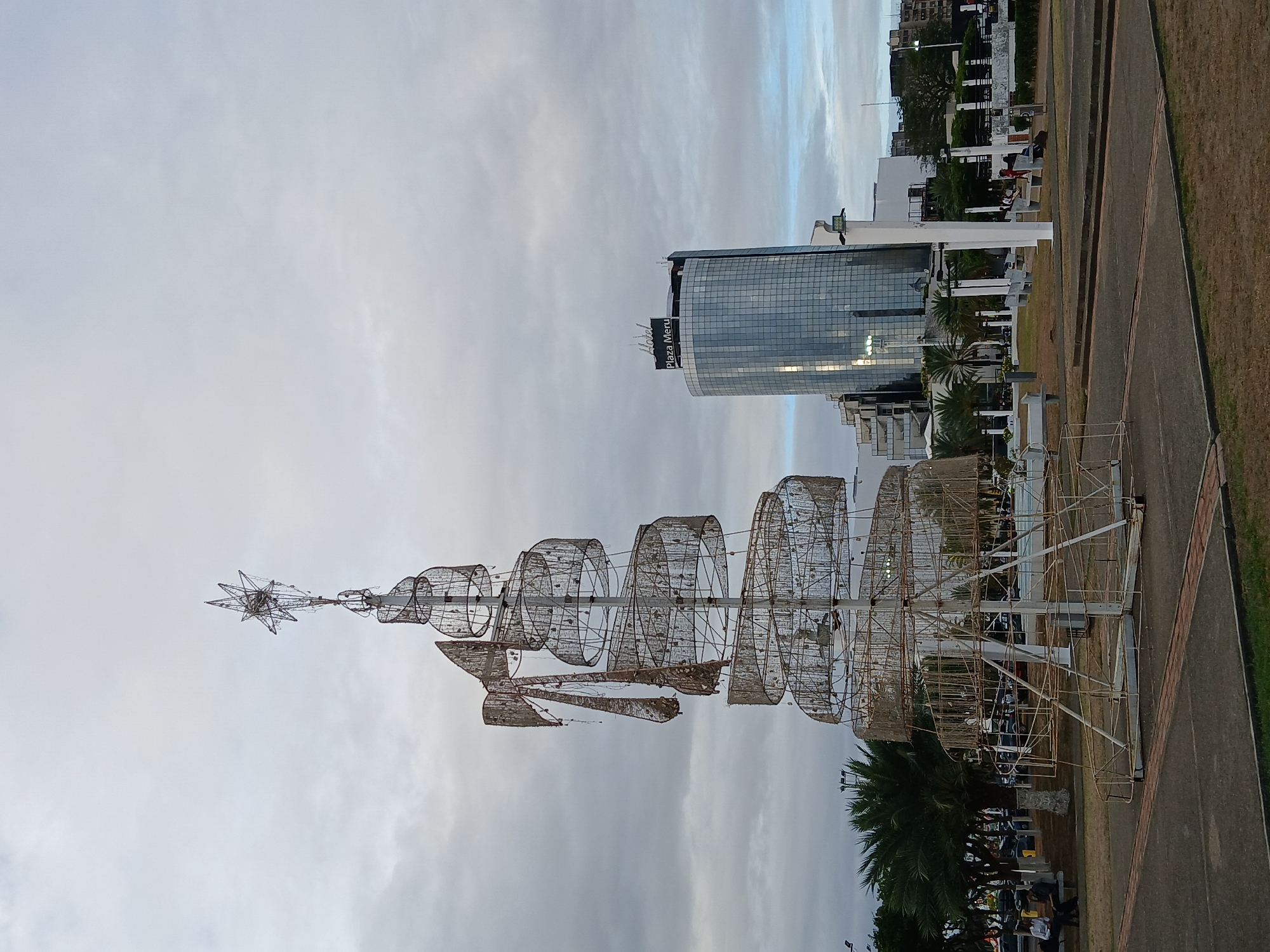 The image depicts an urban park or plaza in Ciudad Guayana, Venezuela, with the Hotel Plaza Meru prominently visible in the background.
In the foreground, a large, free-standing, skeletal Christmas tree structure is centered. It is constructed from a central pole with spiraling, weathered metal bands that form the conical shape of a tree, topped by a star ornament. A large, deteriorated metal bow adorns the upper left side of the structure. The base consists of a square concrete slab.
In the midground to the right, the Hotel Plaza Meru stands as a modern, multi-story building with a reflective, grid-patterned glass facade. The "Hotel Plaza Meru" sign is visible on its rooftop. To the left and behind the tree structure, a line of palm trees and other green foliage separates the park from a street with parked vehicles.
The park area features dry grass and dirt patches intersected by concrete pathways with brick accents. Several white public benches and pillars are scattered throughout the midground. Multiple individuals are present: two males are seated on a bench under palm trees on the far left, one in a white shirt and dark trousers, the other in dark attire. Further to the right, near the white pillars, at least five other individuals are visible, seated or standing on benches and pathways.
The sky is overcast with a uniform layer of light grey clouds, indicating diffuse light conditions. No specific interactions between individuals are discernible.