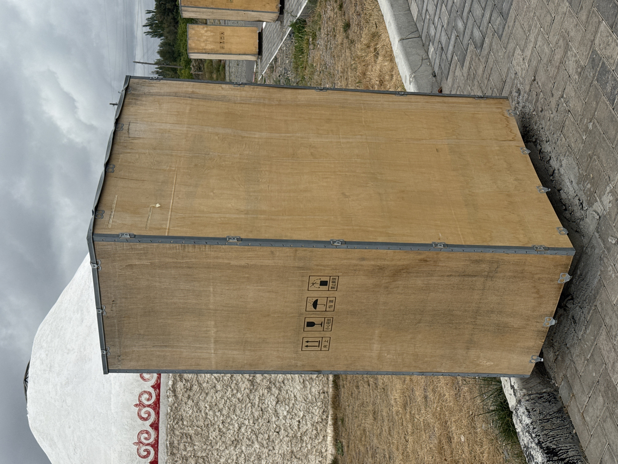In Balykchy, Kyrgyzstan, a large wooden shipping crate stands on a paved area next to dry grass. The crate is tall and rectangular with metal banding and visible hinges. To the left of the crate, a portion of a white yurt with red decorative trim is visible, suggesting a traditional nomadic dwelling. Behind the crate, several other similar crates are lined up, and beyond them, a line of trees and utility poles can be seen against a cloudy sky. The overall environment appears to be an outdoor area, possibly a staging or storage zone, with a mix of natural and constructed elements.