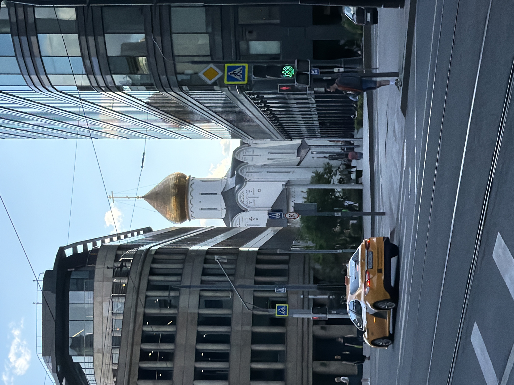 The image shows a street scene in an urban environment. The photograph is composed of modern architecture juxtaposed with a Russian Orthodox church topped with a golden dome, suggesting the location may be in Russia, specifically Moscow. The church is centered in the frame, nestled between tall buildings on either side. Pedestrian crossing signs are visible near the buildings. A yellow taxi is present on the street, adding to the city's ambiance. One pedestrian can be seen walking across the crosswalk. The street also has tram tracks. The sky is partially visible, suggesting it is daytime.