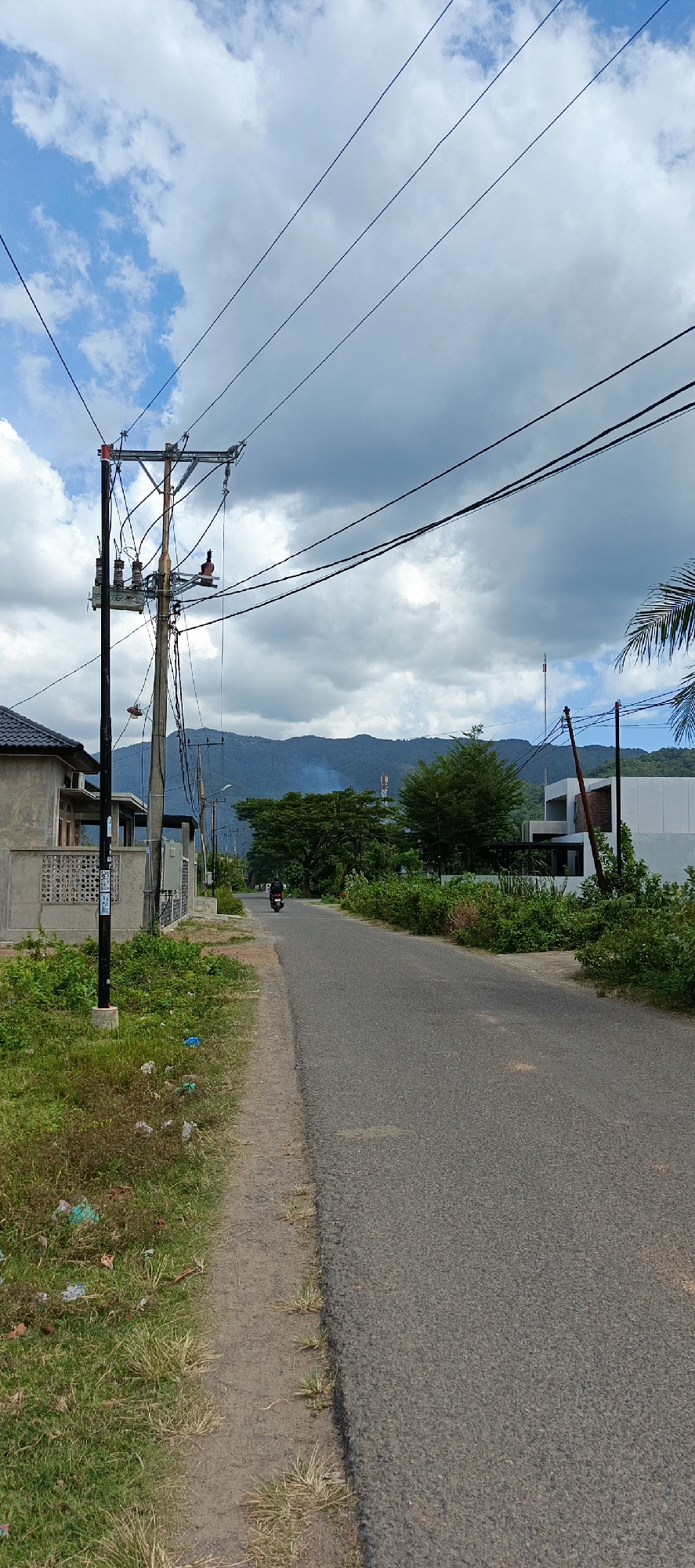 The image depicts a scene on a road in Pashiebeutong, Indonesia. A paved road runs from the foreground into the distance, with a dirt shoulder on the left. The left side of the road is bordered by a grassy area with some scattered litter and a house with a gray facade and a tiled roof. Further down the road, there are several power poles with complex wiring systems. On the right side of the road, there is dense green vegetation and a modern white building with dark window frames. In the background, a line of lush green mountains is visible under a cloudy sky with patches of blue. A single person is riding a motorcycle on the road in the middle distance. The overall environment suggests a semi-urban or suburban setting in a tropical or sub-tropical region.