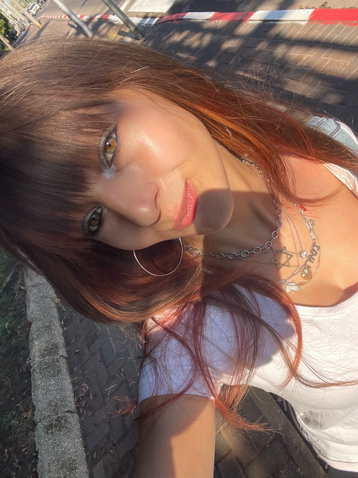The image shows a young woman with auburn hair and light green eyes, wearing a white t-shirt and multiple necklaces, including one with a Star of David pendant. She is outdoors, with a paved area and some greenery visible in the background. The lighting suggests it is daytime, possibly late afternoon due to the long shadows cast on the ground. The background elements, including the red and white striped curb and the paving stones, are consistent with a street or public walkway in an urban environment. While the specific location within Raanana, Israel is not definitively identifiable from the visual information alone, the overall context suggests an outdoor public space in a city.