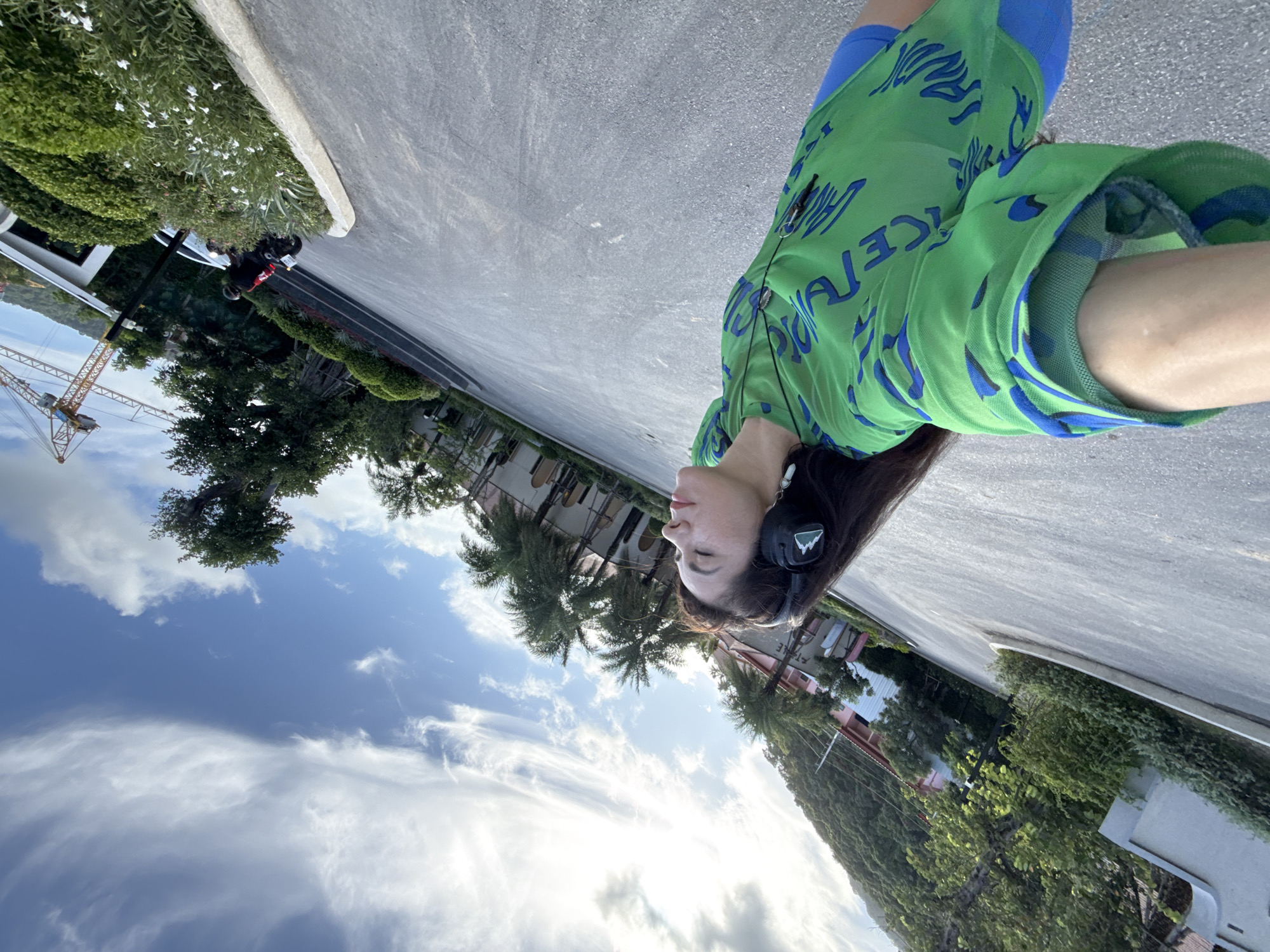 A woman wearing headphones and a green, blue-patterned shirt is standing on an asphalt road in Karon, Thailand. She is looking up at the sky, which is blue with scattered clouds. In the background, there are palm trees, buildings, and a construction crane. A person on a motorcycle is visible in the distance. The sun is shining, creating bright highlights.