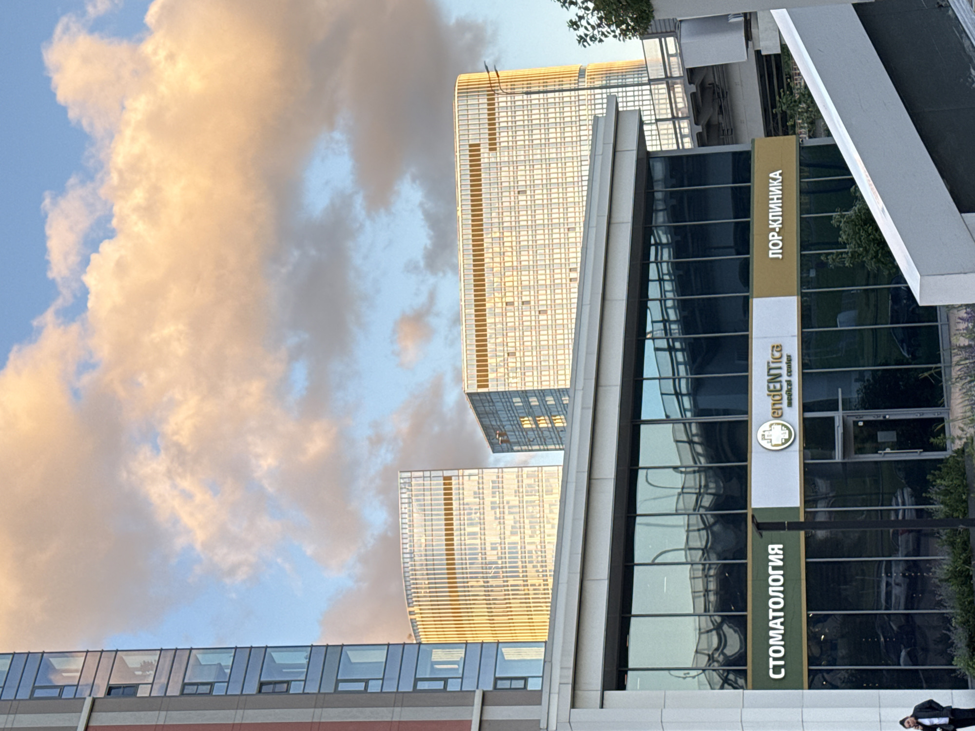 The image captures a modern urban scene in Moscow, Russia, focusing on a contemporary building with glass facades. In the foreground, a low-rise structure houses two medical clinics, indicated by signage. The left sign reads "СТОМАТОЛОГИЯ" (Dentistry) in white letters on a dark green background. To its right, a sign with a green circular logo containing a cross and the word "endENTica" appears in white text, with "medical center" below it. Further to the right, another sign reads "ЛОР-КЛИНИКА" (ENT Clinic) in white letters on a light brown background. Large glass windows dominate the lower building, reflecting the sky and surrounding architecture.
Behind the clinics, two taller, modern buildings with grid-like facades, also featuring extensive glass, are visible. These buildings are illuminated by the warm light of the setting sun, casting a golden glow on their surfaces and the clouds above. The sky is a mix of blue and soft, warm-toned clouds, suggesting it is either sunrise or sunset. A person in dark clothing and a white jacket is visible on the left side of the foreground, standing near the entrance. The overall impression is one of a well-developed urban environment with modern infrastructure.