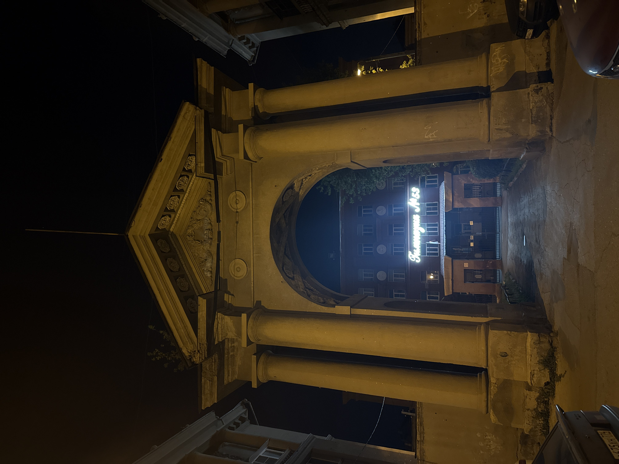 The image depicts a grand, illuminated archway at night. The archway is made of stone and features two large columns. Above the arch, there is a pediment with decorative carvings. Through the arch, a brick building is visible with numerous windows. A neon sign on the building reads "Гимназия №55", indicating it is Gymnasium No. 55. The area in front of the archway is paved and appears to be a courtyard or street. The lighting suggests it is nighttime in an urban environment. The location is identified as Magnitogorsk, Russia.