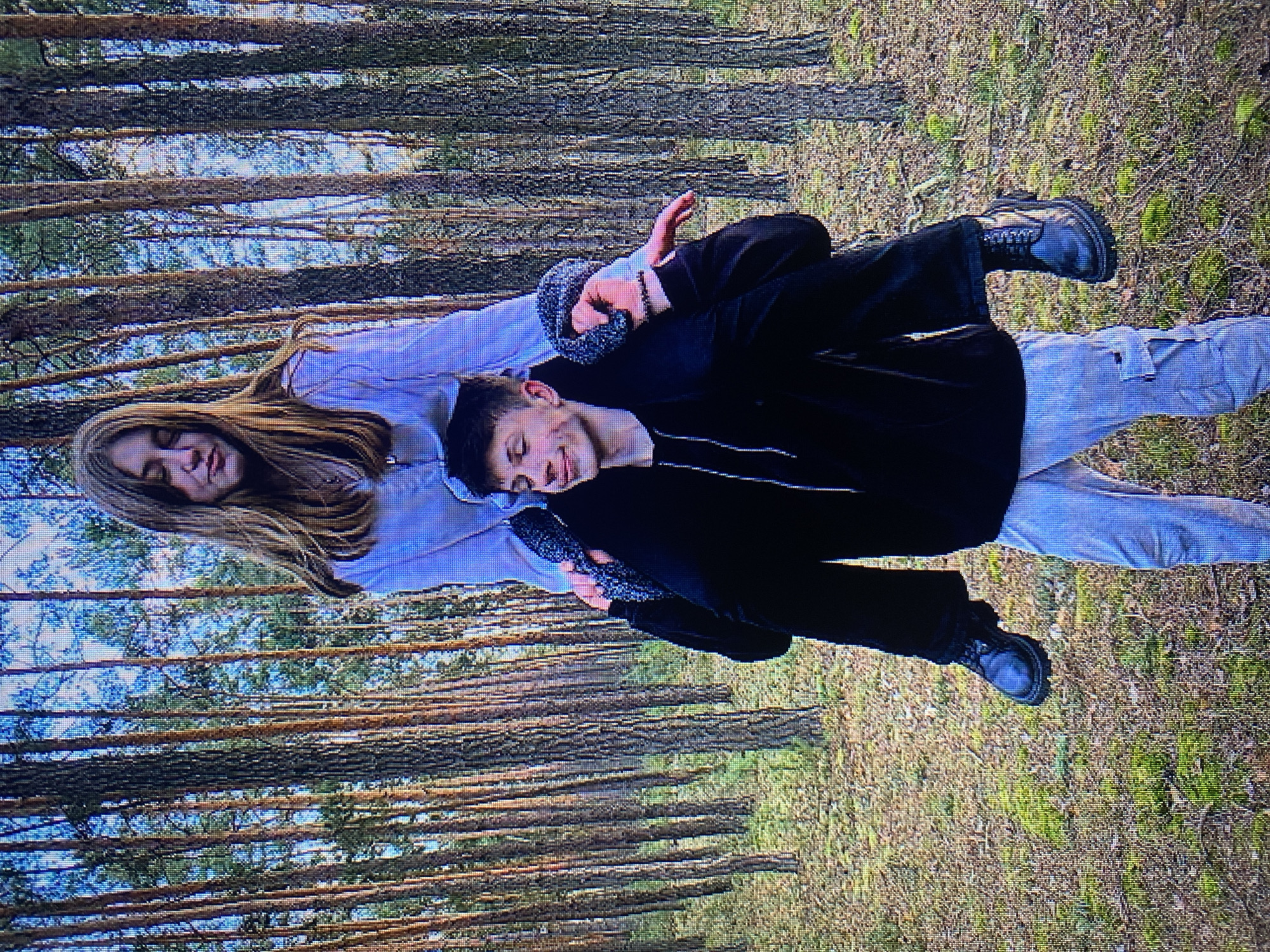 The image depicts two individuals in a pine forest, likely located on the outskirts of Poznan, Poland, given the vegetation and terrain consistent with areas surrounding the city. A young male is carrying a young female on his shoulders. He is wearing a black hoodie and light grey cargo pants, with black lace-up boots. The female, with long blonde hair, is wearing a light blue blouse and is holding onto the male's shoulders. The background consists of numerous tall pine trees with a forest floor covered in pine needles and moss. The lighting suggests it is daytime, possibly late afternoon, with sunlight filtering through the trees.