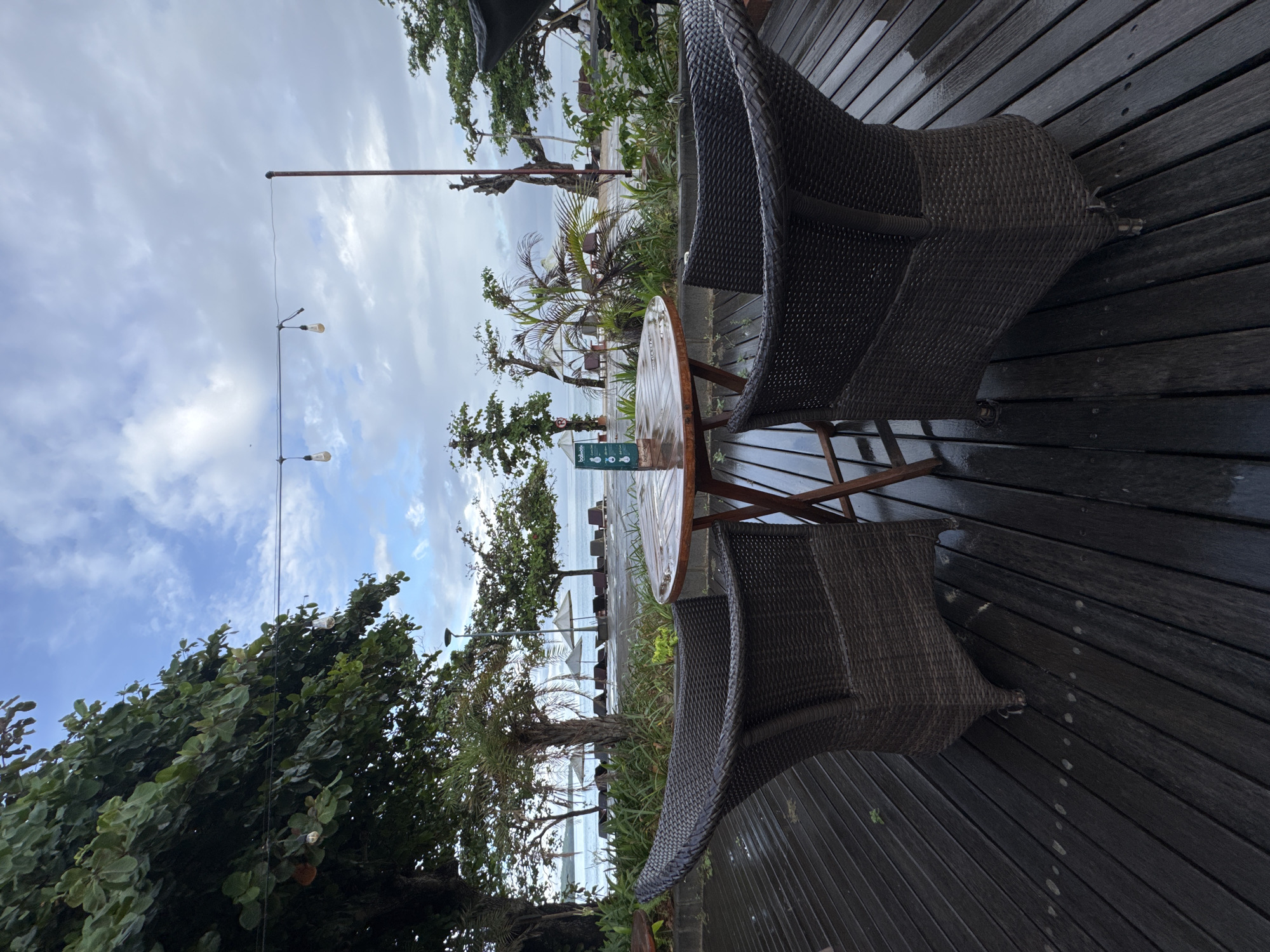 This image shows a rain-slicked outdoor dining area in Kuta Selatan, Indonesia. Two wicker chairs flank a small, round wooden table on a dark wooden deck. Beyond the seating area, vegetation includes trees and palm fronds. In the distance, the sea is visible under a cloudy sky. A string of lights hangs overhead.
