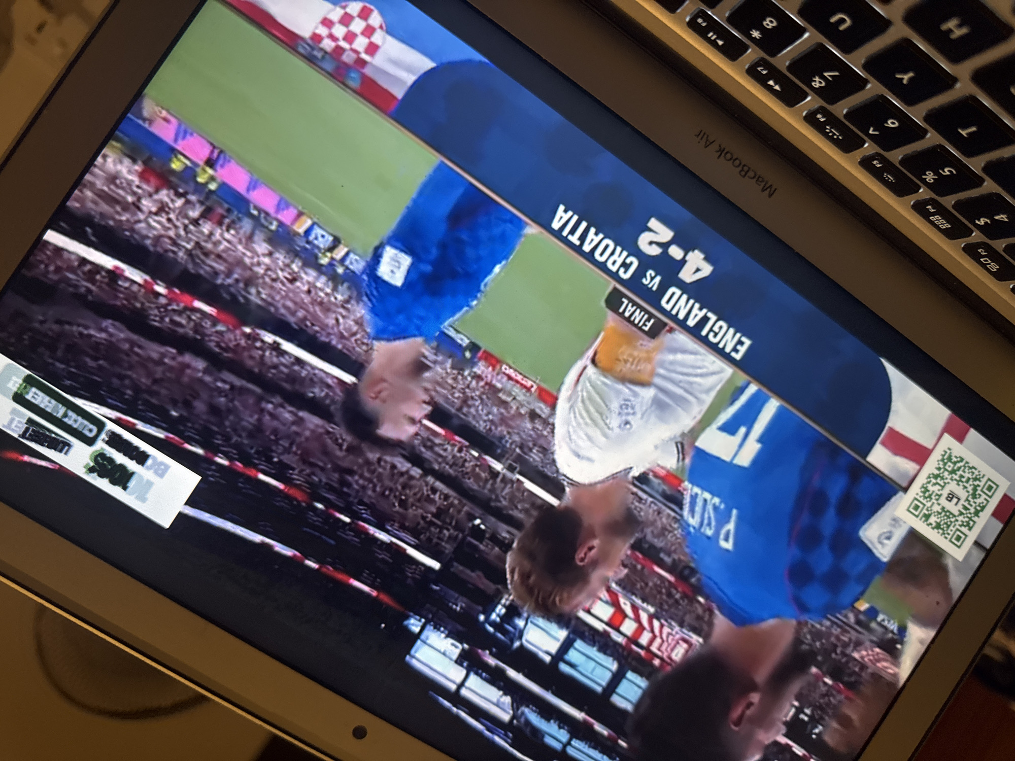 The image shows a laptop screen displaying a soccer match. Two soccer players in blue and white uniforms are visible on the field, with a crowd in the background. A score graphic reads "ENGLAND vs CROATIA 4-2". There is a QR code in the bottom left corner of the screen. The laptop appears to be a MacBook Air. The context of Kufena, Nigeria is not present in the image.