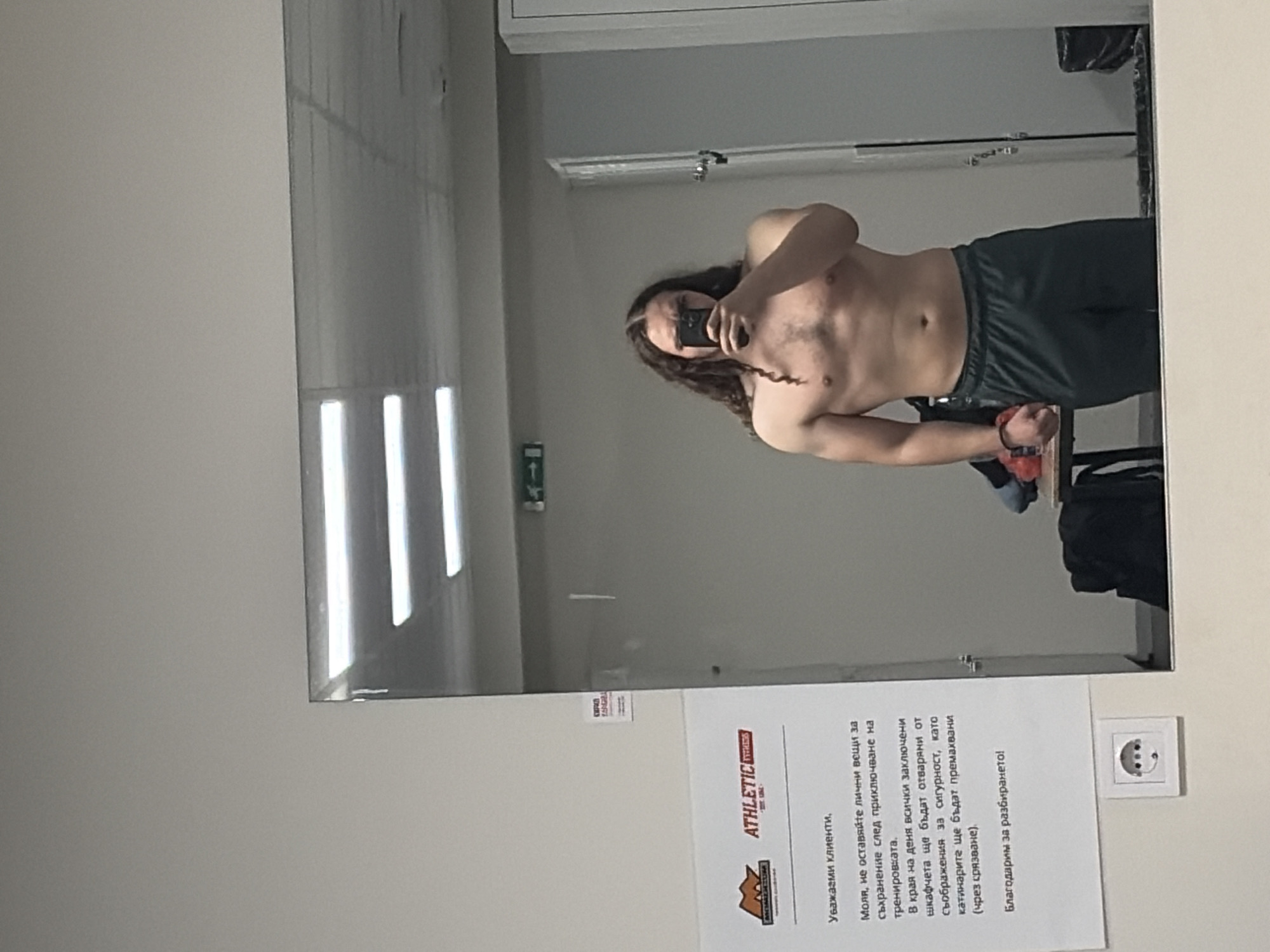 A man with long curly hair is taking a shirtless selfie in a mirror inside what appears to be a gym or fitness center in Plovdiv, Bulgaria. He is wearing dark green shorts and a black fitness tracker on his left wrist. In the background, gym equipment and personal belongings are visible. Affixed to the wall adjacent to the mirror is a sign in Bulgarian, presumably a notice to patrons regarding locker usage and security. An electrical outlet is also visible near the sign. The gym seems modern with overhead fluorescent lighting.