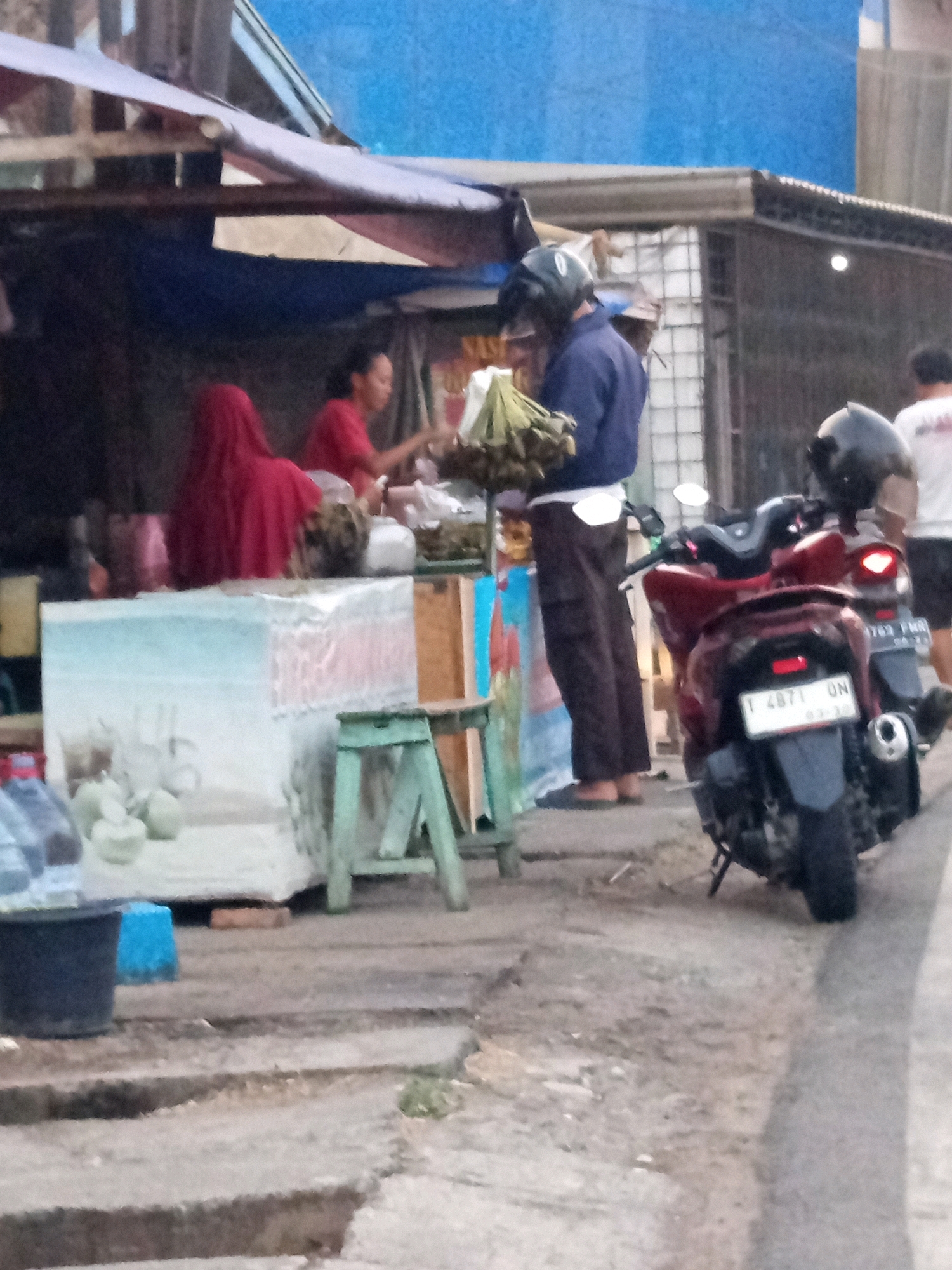 The image depicts a street scene in Karawang, Indonesia, at what appears to be a roadside stall. In the foreground, a red scooter is parked. A person wearing a helmet and a blue jacket is standing near a stall, holding a bunch of what looks like bananas. To the left, a person wearing a red hijab and a red shirt is attending to the stall. Several coconuts are visible on a display in front of the stall, along with bottles of liquid. The stall is covered by a blue tarp and an awning. In the background, other individuals are visible, as well as buildings and a blue tarp. The overall environment suggests a typical urban street setting with commercial activity.