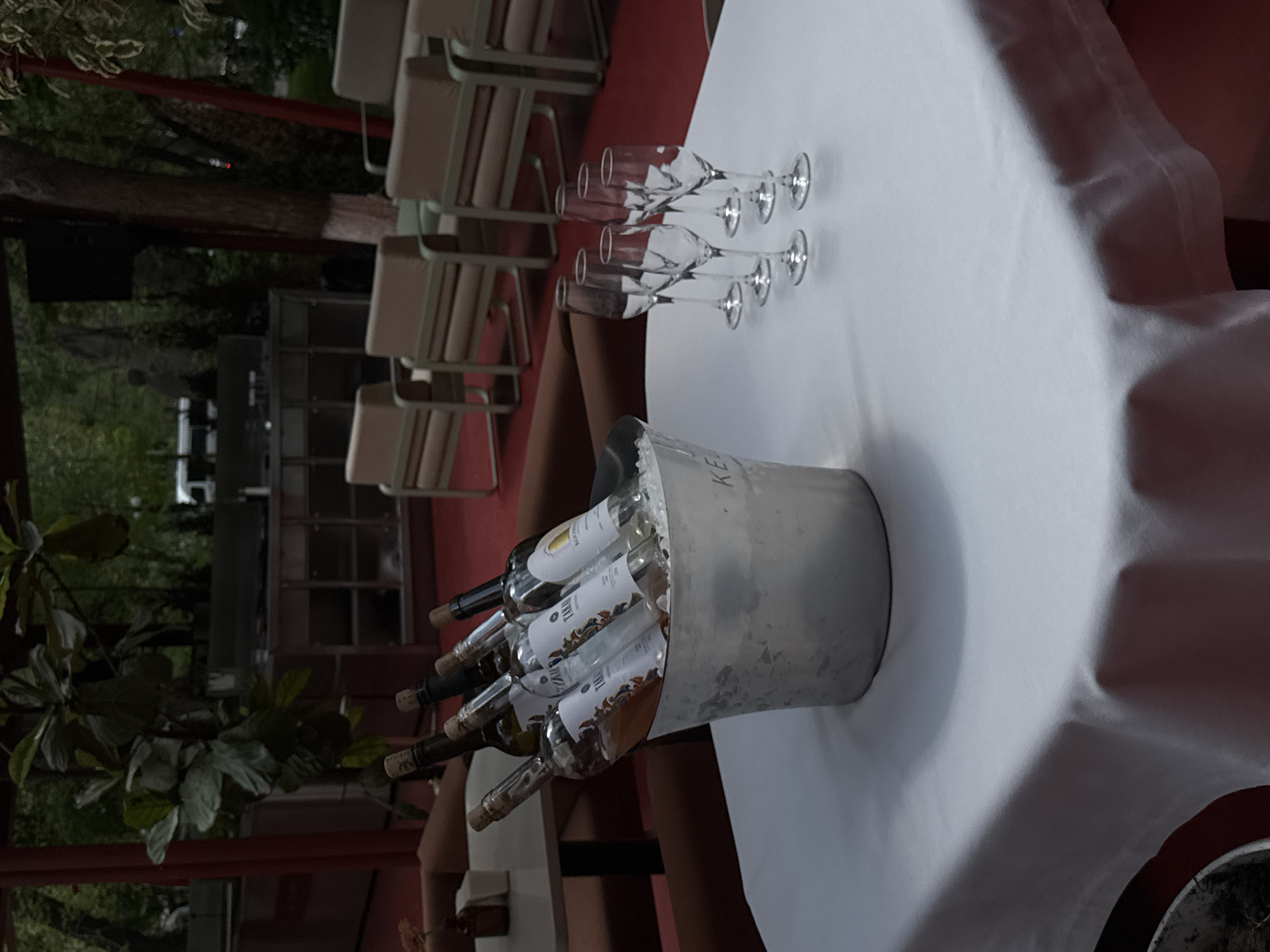 This image depicts an outdoor or semi-outdoor event setup, likely a restaurant patio or garden located in Yerevan, Armenia. The primary focus is a white-clothed table, elegantly prepared for a social gathering.
On the left side of the table, a silver-colored metal ice bucket is prominently placed, filled with several bottles of wine, predominantly white varieties. Some bottle labels are partially visible; discernible text includes "TALIS" or "TALI" on multiple bottles, and "BIANCA" on one label. The letters "KE" can also be faintly seen on the side of the ice bucket, possibly part of a brand or venue name. To the right of the ice bucket, seven empty champagne flutes or wine glasses are neatly arranged in two rows on the white tablecloth, poised for use.
In the background, a series of light-colored, possibly beige, chairs with metallic frames are visible, suggesting an area prepared for guests. Lush green foliage from various plants and trees creates a natural backdrop, indicating an open-air environment. A striking red surface, possibly a floor or decorative wall element, adds a vibrant touch to the scene, while other indistinct, darker architectural structures can be seen further in the distance.
The lighting in the image appears soft and diffused, hinting at a time of day like late afternoon, early evening, or an overcast day, contributing to a calm and inviting atmosphere. While no people are present, the meticulous arrangement of wine, chilled in an ice bucket, and the array of glasses strongly suggest an imminent social event, such as a wine tasting, celebration, or a dinner service. The overall impression is one of quiet anticipation for a pleasant gathering.